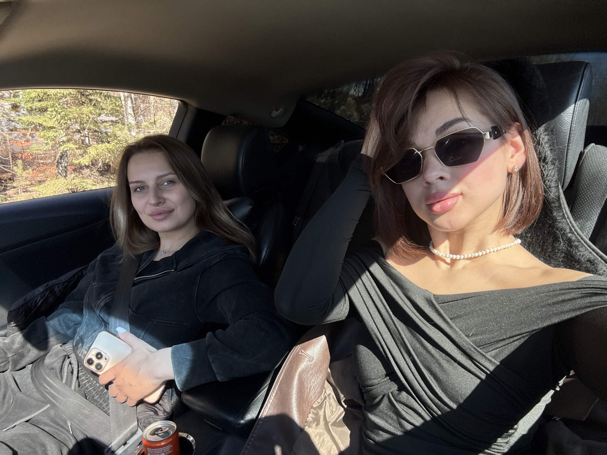 Two women are inside a car. The woman on the left is wearing a dark-colored hooded jacket and pants, holding a smartphone, and has a seatbelt on. A can of soda is visible near her. The woman on the right is wearing a black off-the-shoulder top and a pearl necklace, with sunglasses on her face and her right hand up near her head. The interior of the car is dark, and sunlight is streaming in. Outside the car window on the left, trees can be seen. The location is likely in the vicinity of Лобаново, Russia based on the presence of trees typically seen in the countryside.