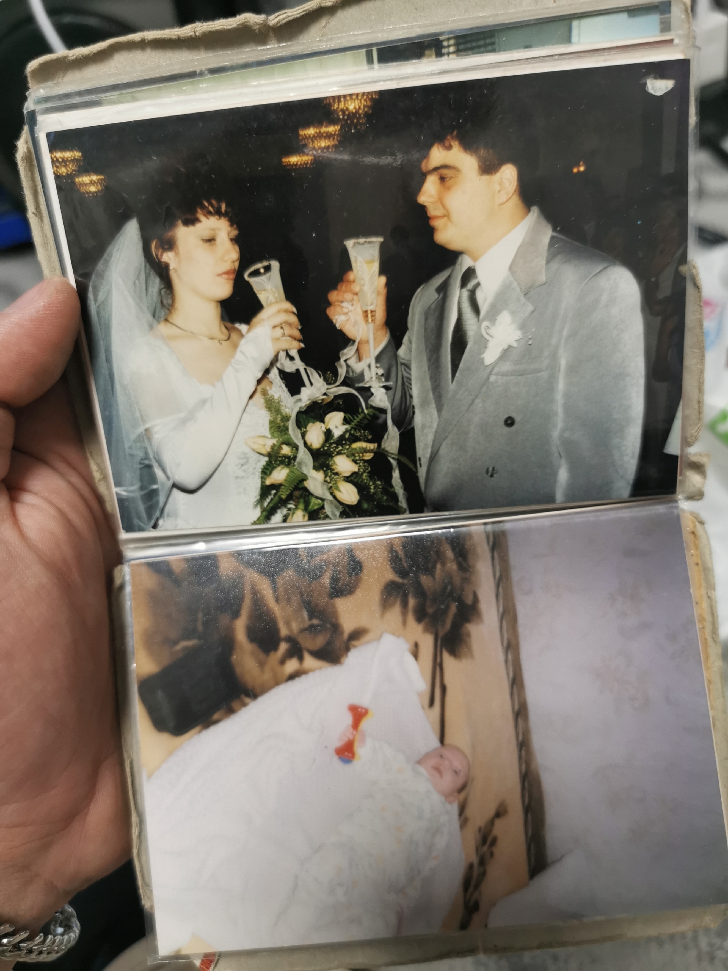 The image shows a hand holding an open photo album. The top photo depicts a wedding. The bride, with dark hair and a veil, is holding a champagne flute. The groom, in a light-colored suit, also holds a flute. They appear to be toasting. The background is dark with chandelier lighting. The bottom photo shows a baby lying in a crib with a patterned blanket. A toy is visible near the baby. The location in Varna, Bulgaria, cannot be determined from the visual information.