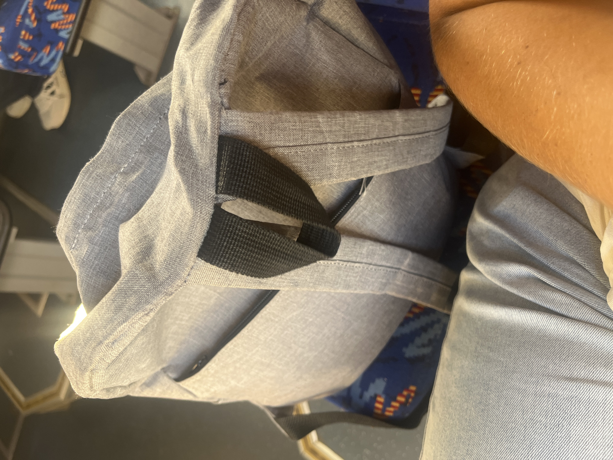 The image shows a close-up view of a gray backpack with black straps, placed on top of a blue seat with a colorful pattern. The backpack appears to be made of canvas or a similar textured fabric. A person's arm, tanned and lightly speckled with what appears to be hair, is visible in the lower right corner of the image. The background is out of focus, but suggests an indoor environment with seating and possibly other passengers or objects. The scene does not provide any specific visual cues to determine the location in the city of Minsk, Belarus.