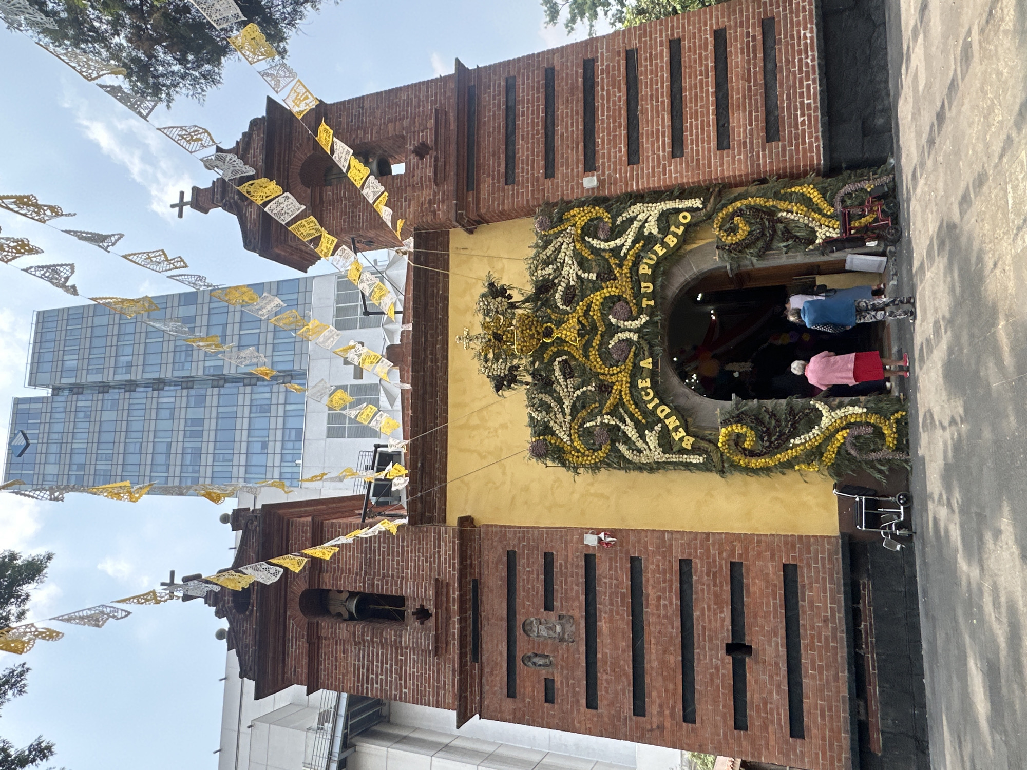 The image depicts a church in Mexico City, Mexico. The church is adorned with floral decorations above the arched entryway, spelling out the words "BENDICE A TU PUEBLO". The church building features brick sections on either side of a yellow facade. Papel picado decorations hang overhead. People are visible entering the church. In the background, a modern high-rise building is visible. The street pavement is visible in the foreground. A walker or wheelchair is near the entrance.