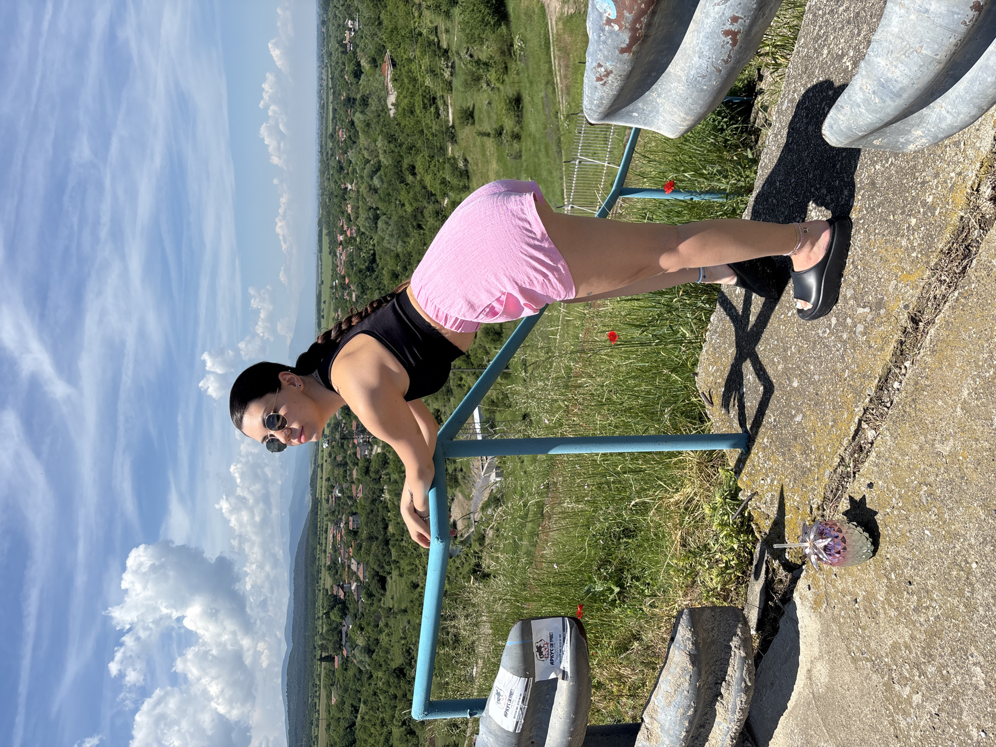 The image, initially rotated 90 degrees clockwise, captures a woman posing on an elevated concrete structure overlooking a vast, scenic landscape. The setting appears to be a sunny day in Ognyanovo, Bulgaria.
The woman, the central subject, is shown in a dynamic pose, leaning forward with her hands resting on a blue metal railing. She wears a black crop top, light pink shorts, and black platform slide sandals. Her dark hair is styled in a long braid, and she is wearing round sunglasses.
Behind the woman, a panoramic view unfolds under a bright blue sky with scattered white clouds. The landscape consists of rolling green hills, dense forests, and agricultural fields. In the distance, several small villages with houses featuring red roofs are visible, nestled among the greenery.
In the foreground, besides the blue railing, there are elements of the concrete structure she is on, along with patches of green grass and a few small red flowers, possibly poppies. A distinctive strawberry-shaped cup with a straw is placed on the concrete surface near her feet. Two large, rusty metal pipes are also visible on the concrete structure. On a cylindrical object near the railing, stickers are affixed. Visible text in Cyrillic reads "АРУССВЕТГМРЕТ" with phone numbers "0877991100" and "0877991160" below it. Another sticker features a stylized dog or wolf logo.
The clear sky and strong shadows indicate bright midday or early afternoon sunlight, suggesting pleasant weather for enjoying the outdoor vista.
