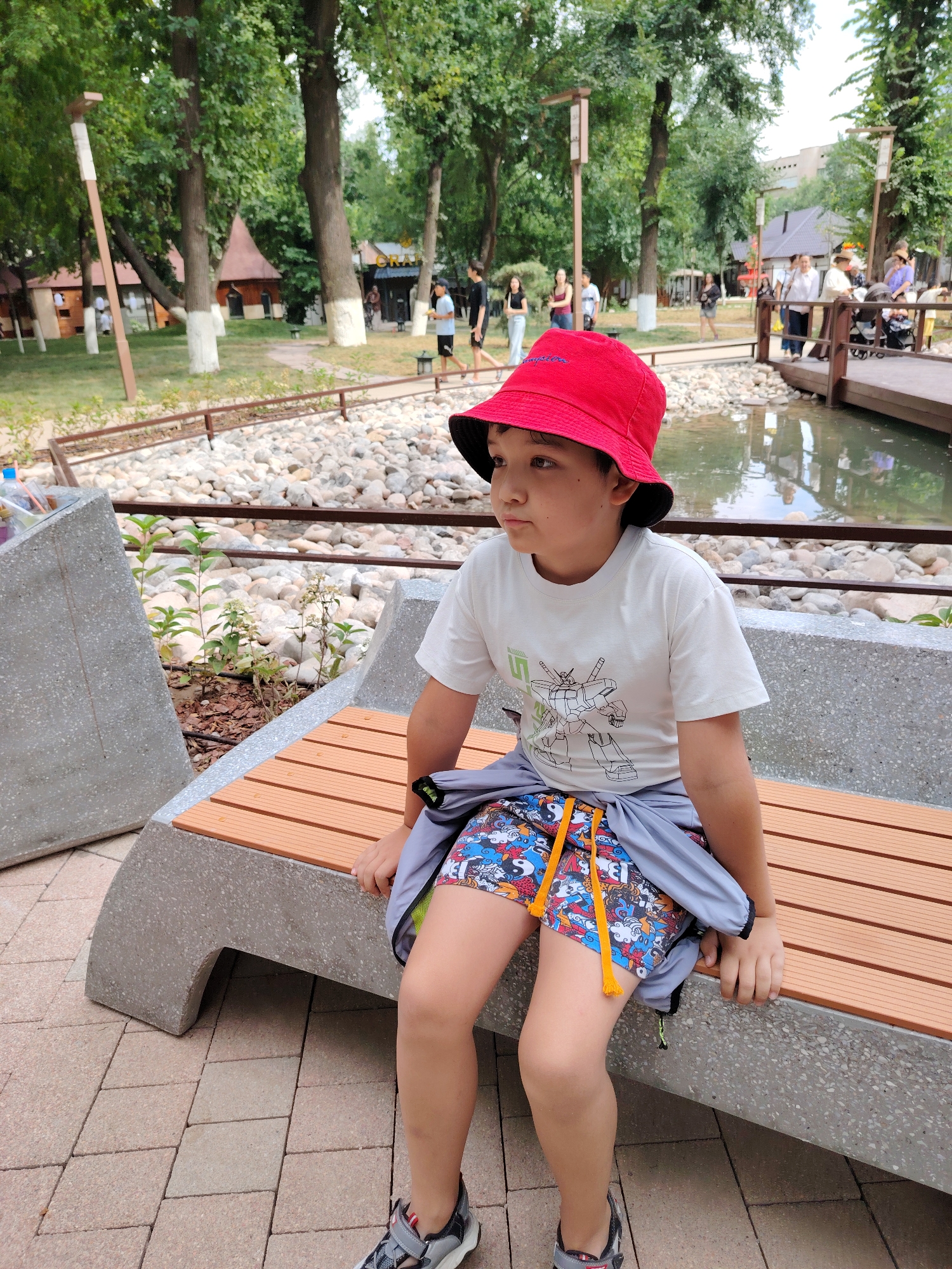 A young boy wearing a red bucket hat and a white t-shirt with a cartoon robot graphic is sitting on a modern bench in an outdoor park. He has on colorful shorts and a grey jacket tied around his waist. The bench is made of concrete and wood. Behind the bench, there is a rocky area and a small body of water with a wooden bridge. In the background, there are trees, buildings, and other people walking around. The ground is paved with interlocking bricks. The scene appears to be during the daytime, under a cloudy sky.