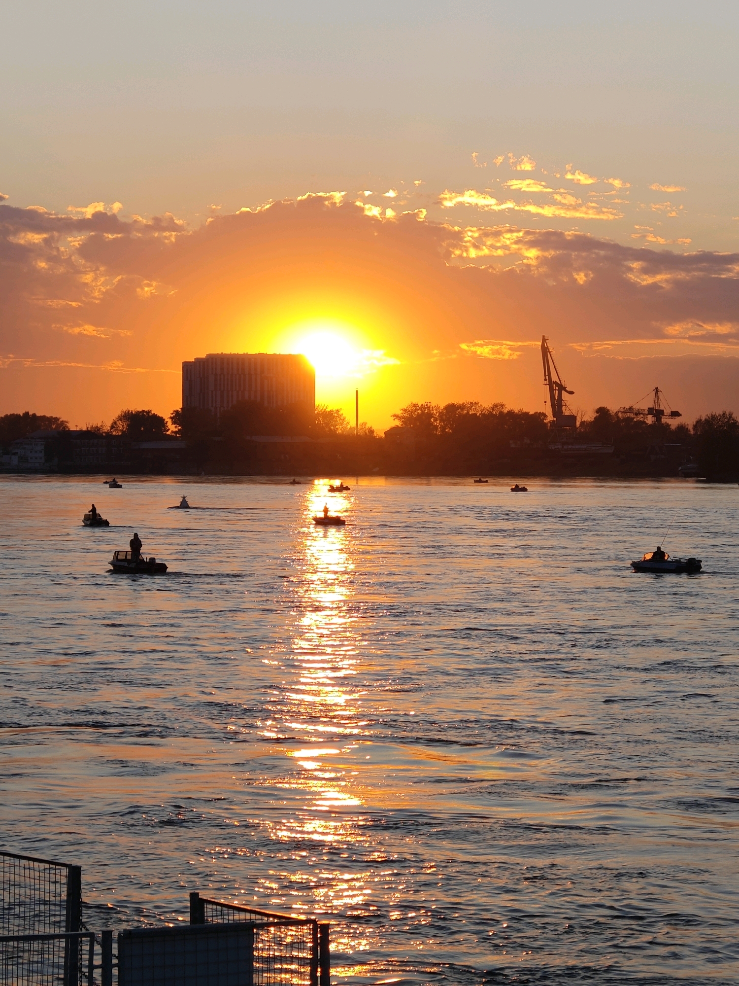 This vertical, eye-level, medium shot captures a sunset over a wide body of water, likely the Angara River in Irkutsk, Russia. The sun, a bright orange orb, is low on the horizon, partially obscured by dramatic clouds that are tinged with orange and yellow. The intense sunlight creates a shimmering reflection on the water's surface, forming a golden path.
Several small motorboats are scattered across the water. In some of these boats, individuals can be seen fishing or simply enjoying the view, their silhouettes outlined against the bright sky. In the background, a large, multi-story building with a dark facade stands on the distant shore, flanked by trees and industrial cranes. The shoreline is lined with structures, suggesting an urban or semi-industrial environment. In the foreground, part of a metallic fence or railing is visible on the left, indicating the vantage point is from a pier or waterfront structure. The overall atmosphere is serene and picturesque, characteristic of a late summer or early autumn evening.