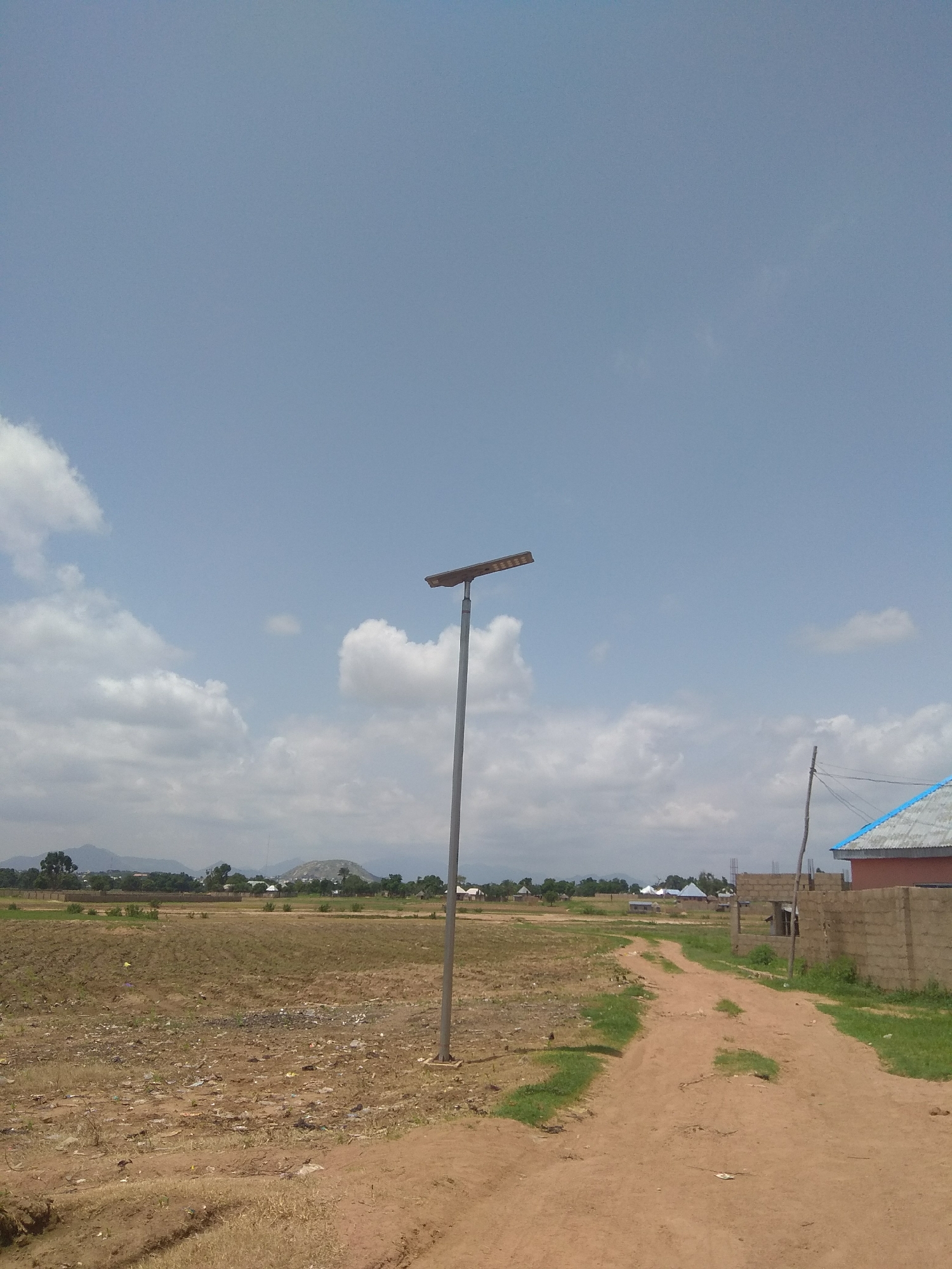 This media file shows an outdoor scene in Jalingo, Nigeria. The primary subject is a solar-powered street lamp standing tall on a dirt path that curves through a rural or developing area. The path is bordered by dry, brown soil and patches of green grass. In the background, there are fields, some appearing recently tilled or uncultivated, with scattered debris. Further in the distance, a small settlement of buildings can be seen, along with trees and rolling hills or mountains under a vast, partly cloudy sky.
To the right of the path, a building constructed with cinder blocks is visible, with a section of a roof covered in blue material. A simple wooden pole with wires is also present near this building. The sky is a clear blue with scattered white clouds, suggesting daytime. The overall impression is one of a dry, possibly hot environment, typical of a rural Nigerian setting undergoing development. No people are visible in the image. The scene appears peaceful and undeveloped. No text is visible.