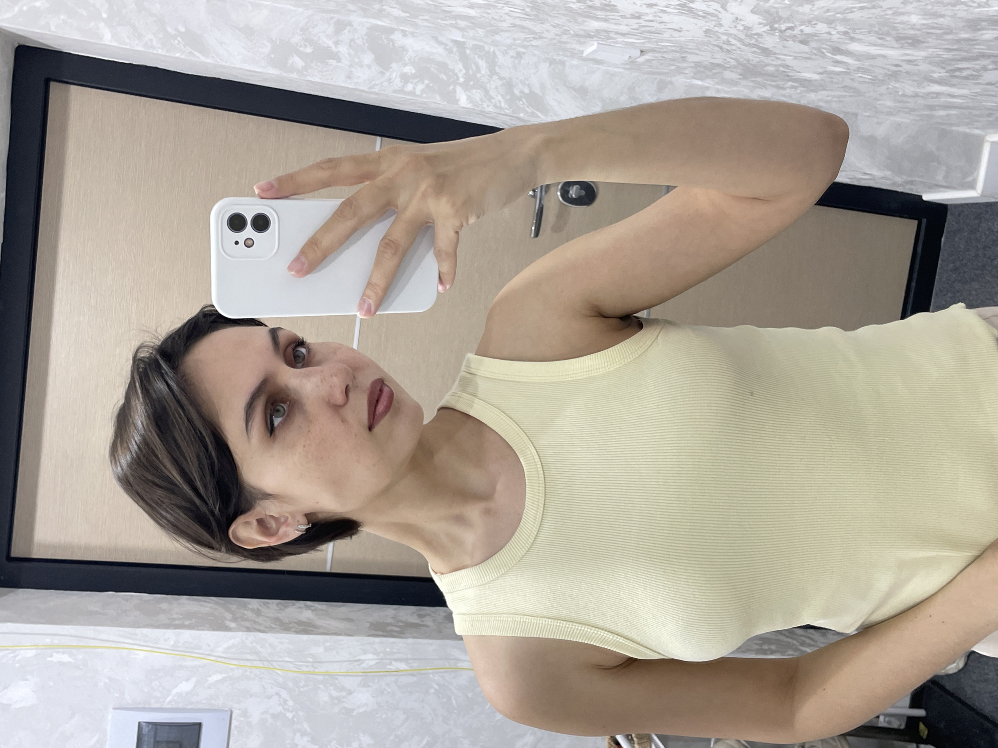 A woman is taking a selfie in a bathroom. She is holding a white smartphone with both hands. She has short brown hair, fair skin with freckles, and is wearing a pale yellow tank top. The background shows a framed mirror and a white marble-patterned wall. A gray floor and a glimpse of a toilet are visible in the lower right corner. A light switch is on the lower left wall. No specific location in Izhevsk, Russia is identifiable.