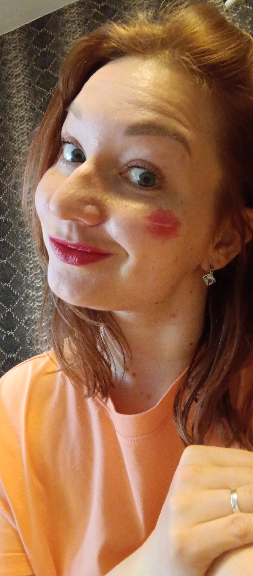 A woman with red hair and fair skin poses indoors. She is wearing an orange t-shirt and has a red lipstick mark on her right cheek. She is also wearing a silver ring on her left ring finger and a silver earring on her right ear. The background is a patterned wall. The location is Unknown, Unknown.