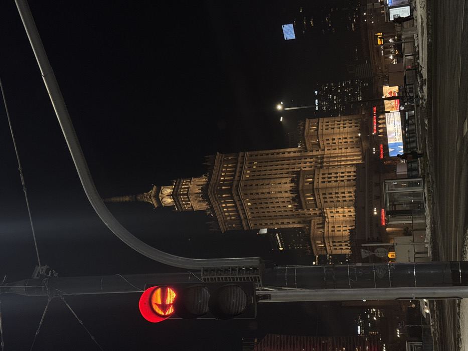 At night, the Palace of Culture and Science in Warsaw is illuminated, towering behind a traffic light displaying a red signal with a whimsical smiling face. Streetlights dot the cityscape. A vertical pole and curved wires intersect the scene. The architecture exhibits intricate detailing, contrasted against the dark sky. Buildings and billboards line the street.