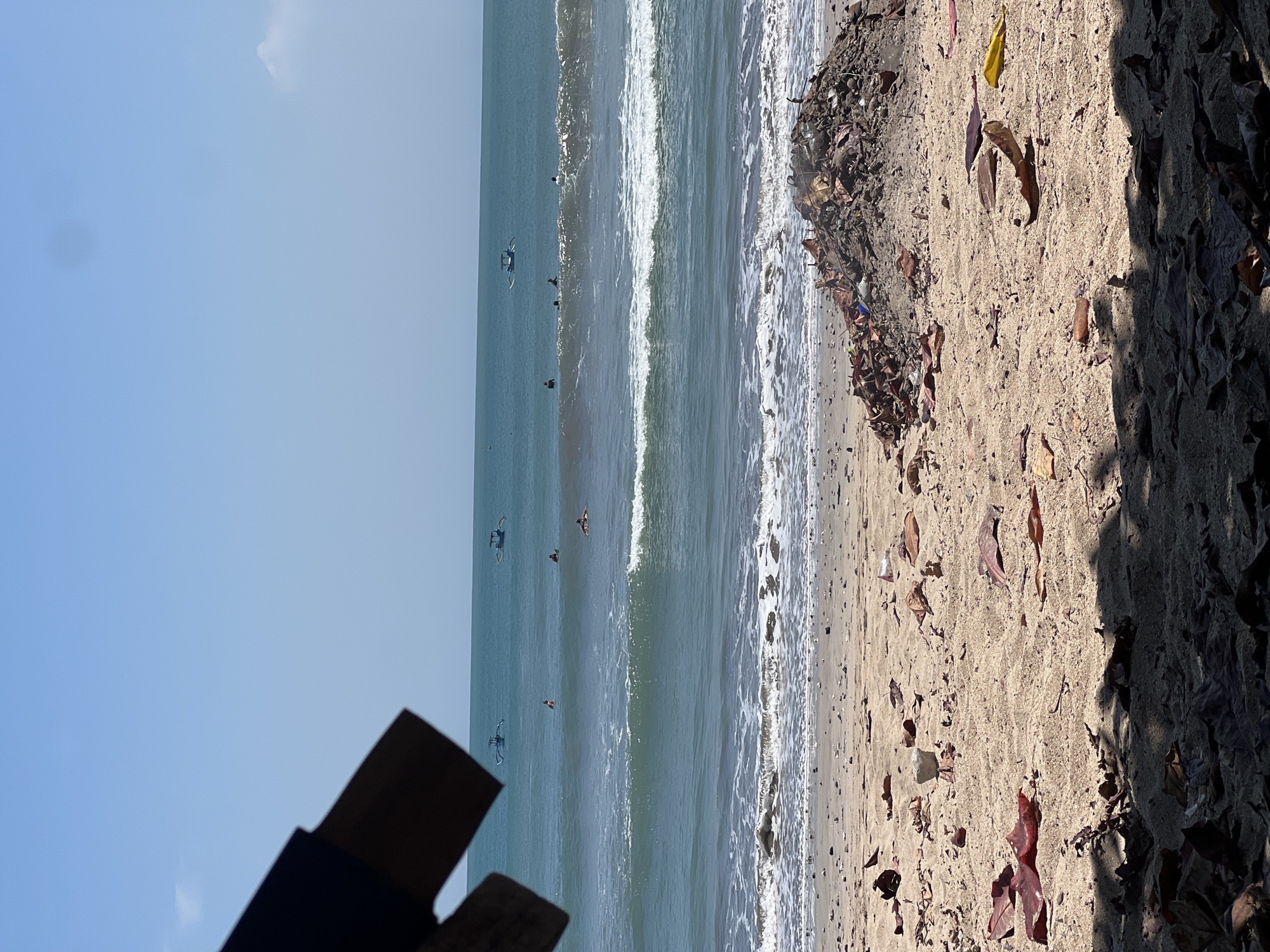 This is a photograph taken on a beach in Kuta, Indonesia. The scene is dominated by the ocean and the sandy shore. In the foreground, the sand is littered with dried leaves and a large pile of debris, including plastic and other trash. Further down the beach, there are shadows cast by foliage. The ocean is a vibrant turquoise, with gentle waves breaking near the shore. Several people are visible in the water, some of whom appear to be surfers. In the distance, two traditional Balinese fishing boats, known as "jukung," are sailing on the calm sea, and the sky is a clear, bright blue. The overall impression is one of a tropical beach scene, marred by the presence of trash.