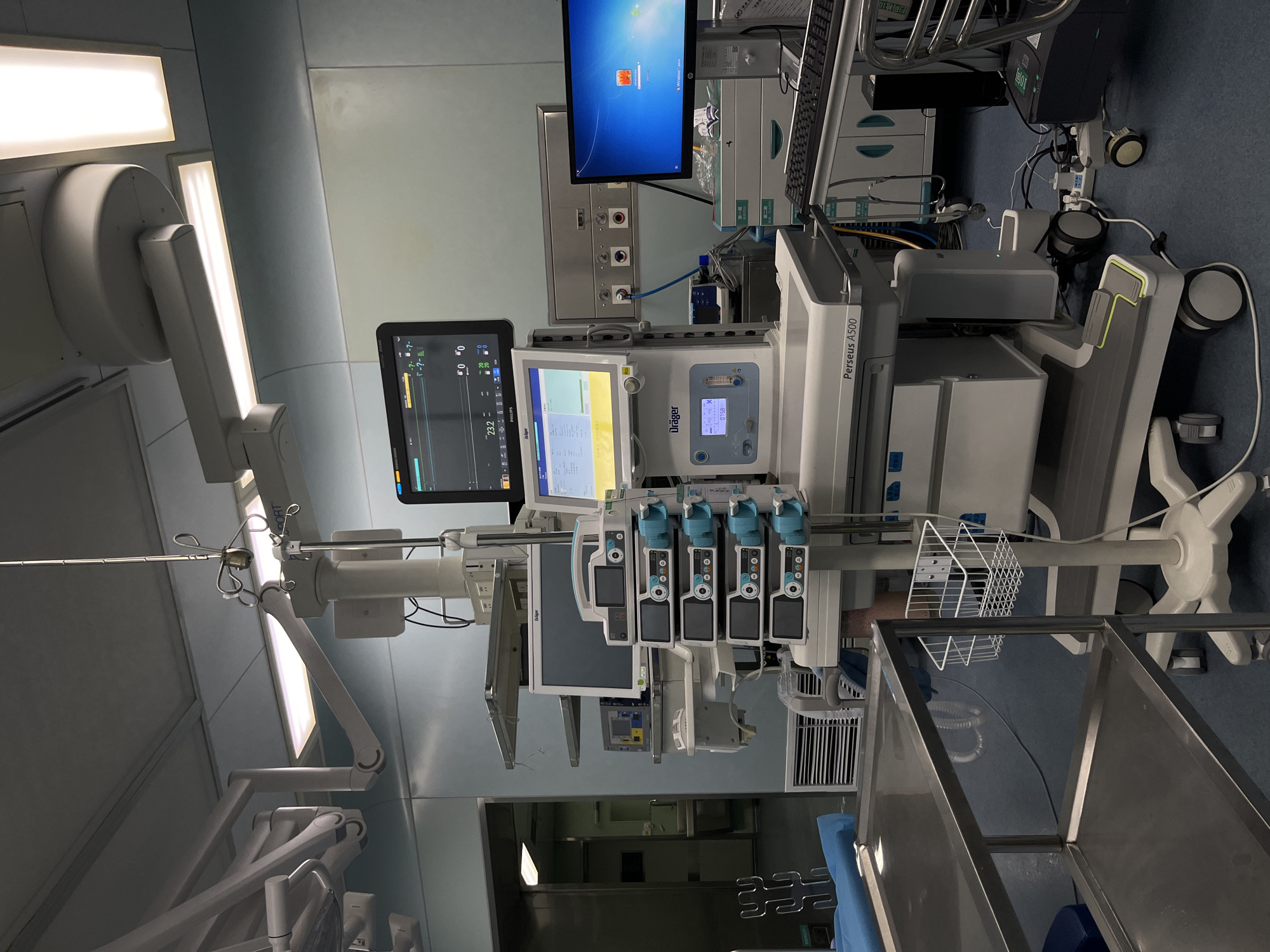 This image depicts a modern operating room, likely in a hospital. Dominating the scene is a Dräger Perseus A500 anesthesia machine with multiple monitors and control panels. The machine is equipped with various medical devices and displays, indicating its function in monitoring and delivering anesthesia. To the left, surgical lights are visible, indicating an operating environment. A Windows monitor is seen in the background along with another piece of equipment possibly used for more vital sign monitoring. In the foreground, there is a stainless steel table, possibly for surgical instruments. The overall environment appears to be clean and well-equipped, consistent with a medical facility. Based on the OCR content "扫码枪10" being in Chinese, and the city of Yuzhong District, China being requested in the prompt, the location is likely in Yuzhong District, China, in an operating room of a medical facility.