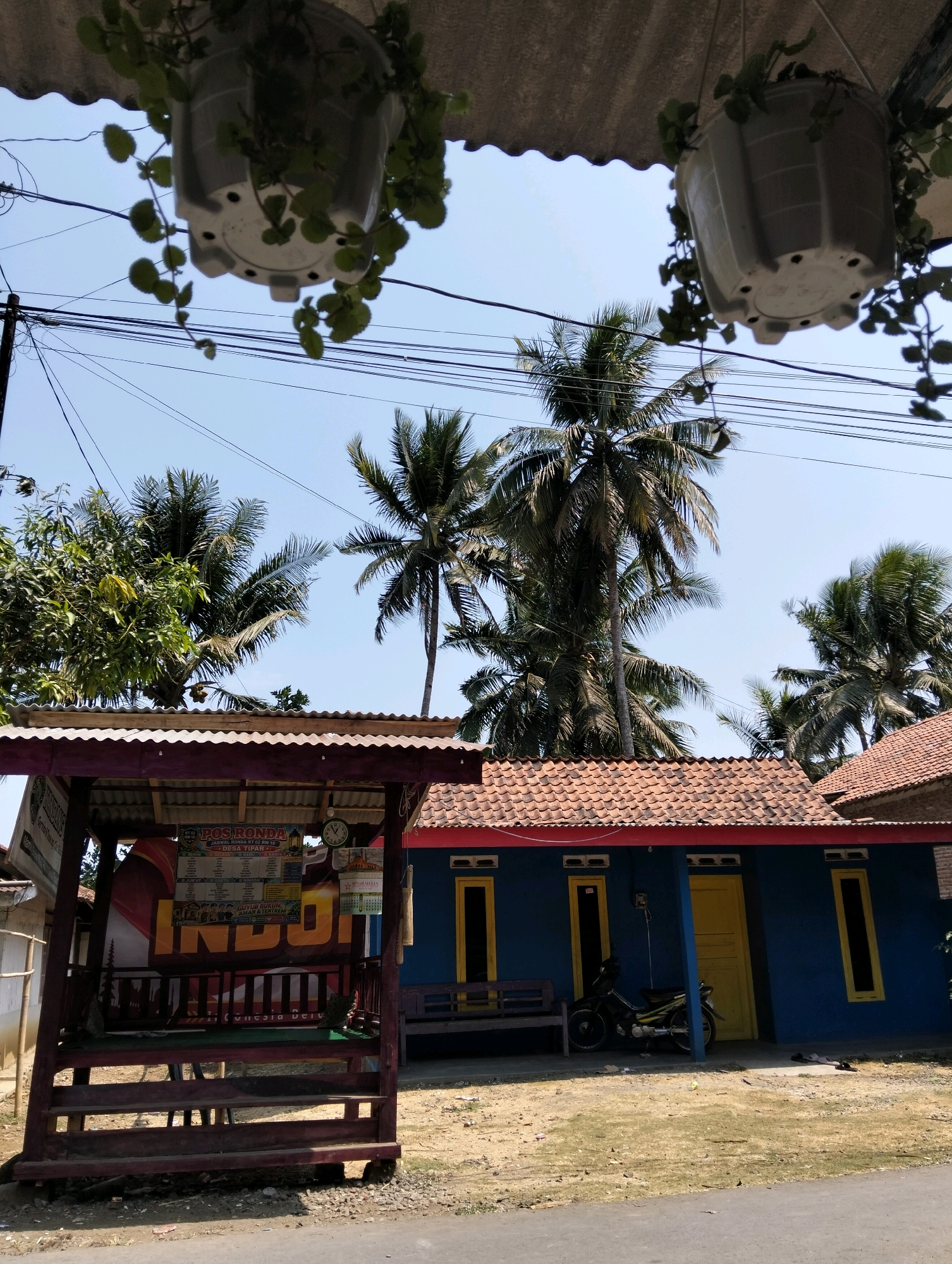 The image depicts a scene in the village of Tipar, Indonesia. In the foreground, a wooden structure with a corrugated metal roof serves as a "POS RONDA" (neighborhood watch post). A banner displaying "INDONESIA" in large letters is visible within the structure, along with a schedule for RT 02 RW 10. To the right, a bright blue house with a red-tiled roof features yellow-framed windows and a yellow door. A parked motorcycle is situated near the entrance. Palm trees and other greenery are abundant in the background, suggesting a tropical environment. The sky is clear and blue. Hanging planters with green foliage are suspended from an overhang above the scene.