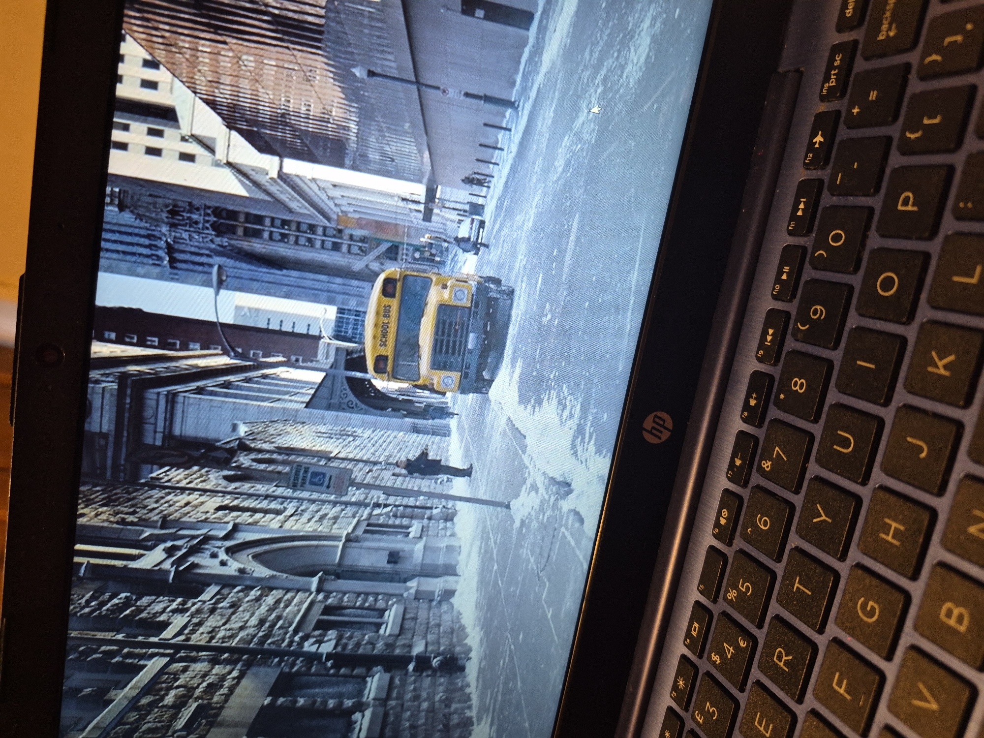 The image displays a laptop screen showing a scene of a yellow school bus parked on a snow-covered city street, lined with old stone buildings. The architecture suggests a European setting, possibly a historic district. A person stands on the sidewalk near a parking sign. The overall environment is wintry and appears to be a still from a movie or video game. The laptop keyboard is visible in the foreground. There is no indication of Sandwell, United Kingdom in the image.