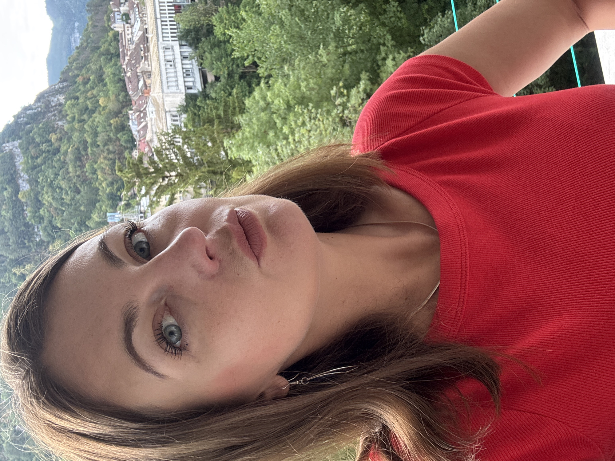 A woman with brown hair and blue eyes is taking a selfie outdoors. She is wearing a red t-shirt and silver earrings. Behind her, a town with red-tiled roofs is nestled at the base of a lush green mountain. The architecture suggests a European setting, likely within the Jura region, possibly the city of Saint-Claude, France, known for its picturesque mountainous landscape and historic buildings. The woman's gaze is directed at the camera, with a slightly pursed expression. The lighting is natural, indicating daylight conditions. The scene appears calm and scenic, with a focus on the woman and the backdrop.