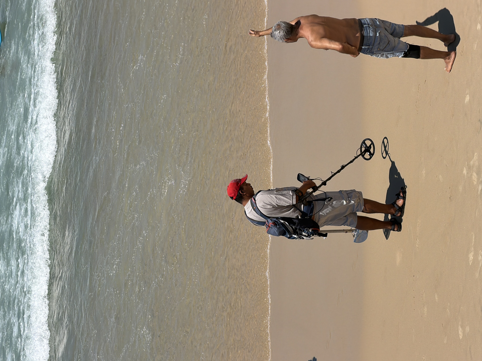 Here is a detailed description of the image:
**Location:** The image appears to be taken at a beach, likely in Tel-Aviv, Israel, considering the Mediterranean coastal environment.
**Environment:** The scene is a sunny day at the beach. The left side shows the Mediterranean Sea with white-capped waves approaching the shore. The beach itself is sandy with some dampness from the water.
**People:** Two men are visible.
* One is shirtless, wearing grey shorts and is standing near the sea with his arm outstretched.
* The other man is wearing a t-shirt, grey shorts, a red cap, and sandals. He has a backpack and is holding a metal detector.
**Objects:**
* Metal detector: The man is using a metal detector, which consists of a control box, a shaft, and a search coil.
* Backpack: The man also has a backpack.
**Actions/Interactions:**
* The man with the metal detector appears to be searching the sand for metal objects.
* The shirtless man is standing near the water's edge with his arm outstretched toward the sea.
**Summary:**
The image captures a sunny day at a beach, possibly in Tel-Aviv, Israel, where a man is using a metal detector to search the sand while another man stands shirtless near the water.