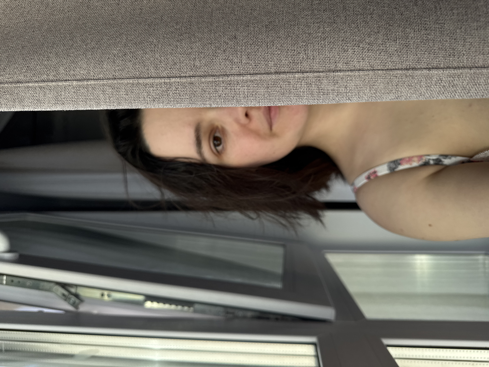 A partial indoor scene in Izhevsk, Russia, features a female individual obscured by a curtain. The subject's right facial profile, encompassing the right eye, nostril, and portion of the mouth, is visible. Her dark brown hair frames this side of her face. Her left shoulder and upper chest are also visible, clad in a white garment with a floral-patterned strap. A light brown, textured fabric curtain with a vertical seam occupies the right half of the frame, concealing the left side of the subject's face and body. To the left of the subject, a white PVC window frame is present, with one pane tilted open inwards, revealing metal hardware. Natural light enters from the window. The subject's gaze is directed forward.