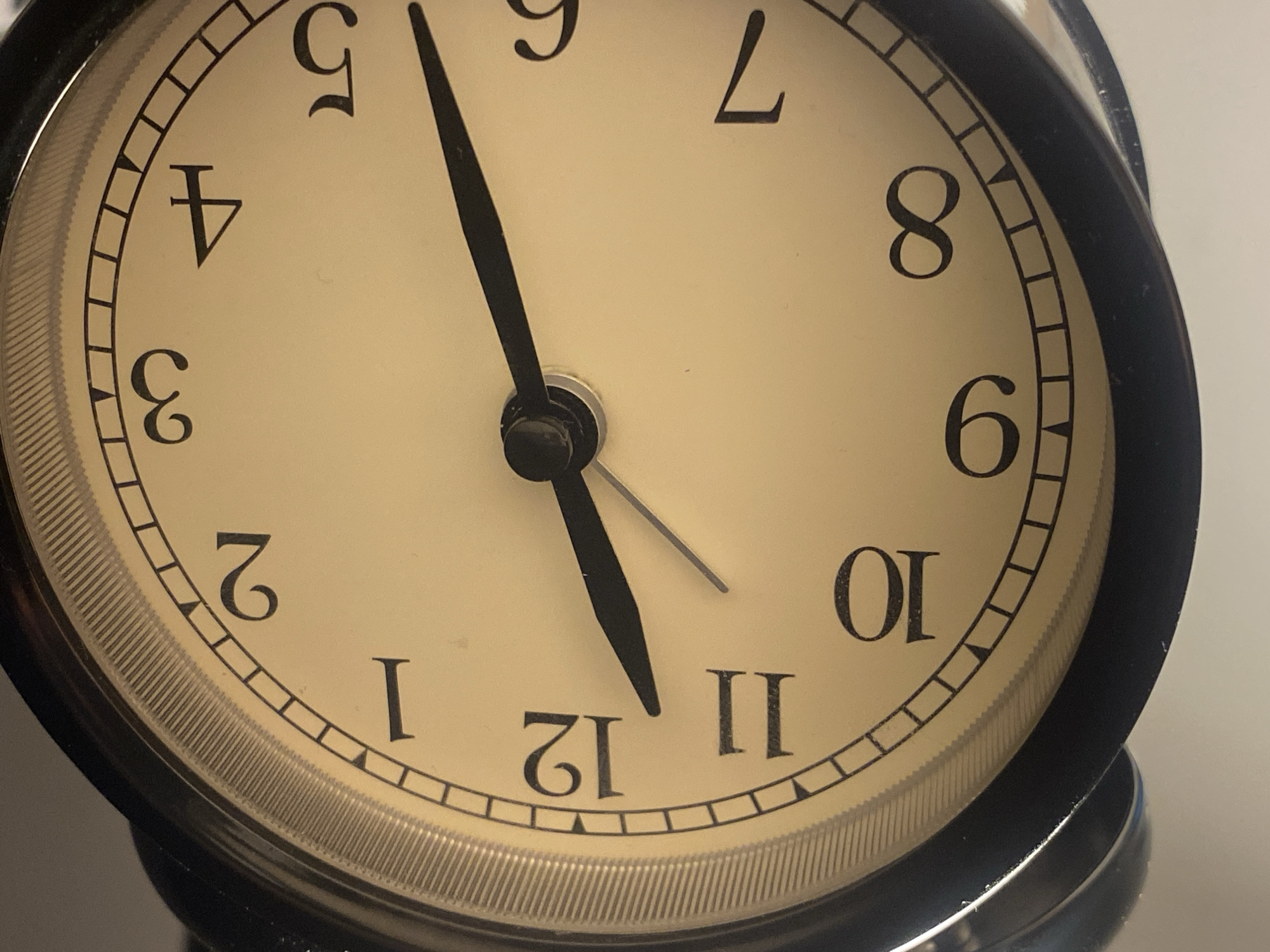 The image shows a close-up of an analog alarm clock with a cream-colored face and black numbers. The clock has black hour and minute hands. The hour hand is pointing just before the 11, and the minute hand is pointing at the 10. The clock is a typical alarm clock design, with a black metal casing. There is no indication of its location in Warsaw, Poland, or any surrounding environment.