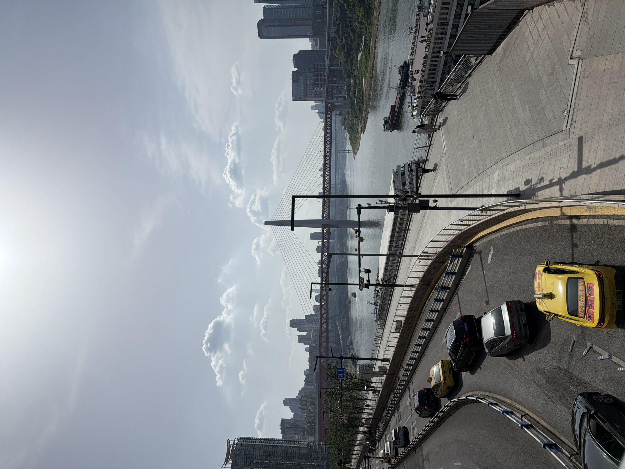 This image captures a vibrant urban scene in Yuzhong District, China, likely during daytime, with bright sunlight and a partially cloudy sky. The perspective is from an elevated viewpoint, looking down and across a city river.
In the foreground, a curved asphalt road occupies the bottom and right side of the frame. Several cars are visible on this road, including prominent yellow taxis with Chinese characters on their rear windows, advertising or taxi service information (e.g., "伴你玩就去逛嘉", "渝A 0TJ350"). Other vehicles are dark-colored sedans, some with visible Chongqing license plates (e.g., "渝B 5X0_0"). The road is bordered by blue and white barriers and features traffic signs, such as a blue sign indicating a right turn for cars and a red circular sign.
To the left of the road, a wide riverside promenade stretches along the riverbank. A few individuals can be seen walking on this paved area. Shadows of lampposts and other structures are cast across the promenade.
Further across, a broad river flows, dotted with various boats and ships. One notable vessel is a large cruise ship with "ELEVATOR CRUISE" visible on its side, and a cargo barge is also present.
Dominating the midground and background is a large cityscape. A distinctive cable-stayed suspension bridge spans the river, connecting the two banks. Both sides of the river are densely packed with numerous high-rise buildings, forming a towering urban skyline under a mix of clear sky and scattered clouds. Sunlight emanates brightly from the upper-left portion of the sky, casting shadows and illuminating the scene.