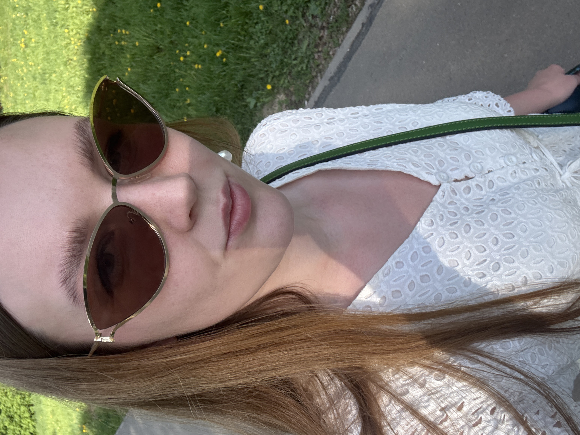 A young fair-skinned woman wearing gold framed sunglasses and a white textured garment with a green strap is pictured from a close, angled perspective. The background features a patch of green grass with small yellow flowers and a grey concrete surface. The location is not determinable from the image.