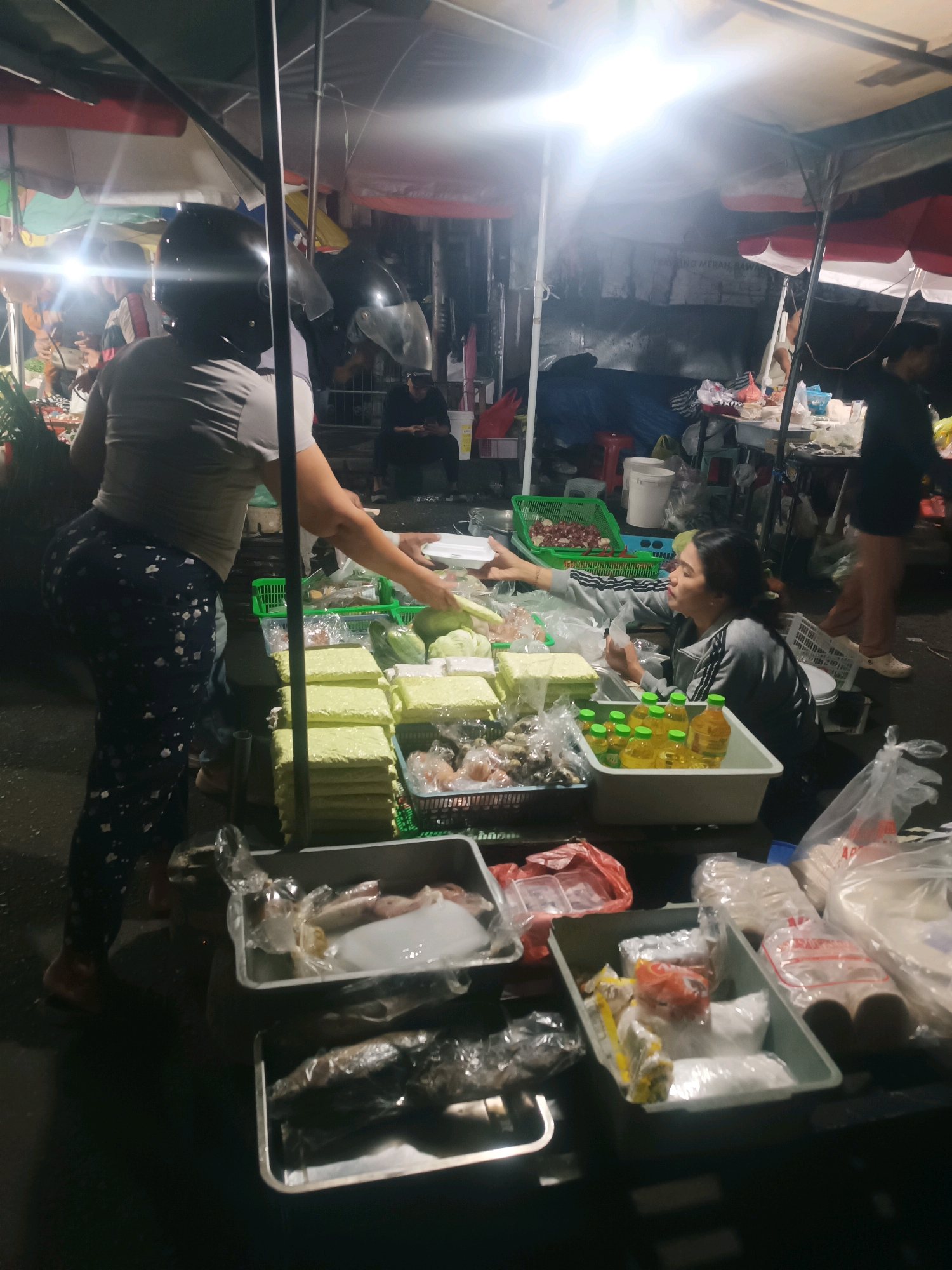 The image depicts a nighttime street scene at a market in Tabanan, Indonesia, likely a food stall or an open-air market. Several individuals are present, engaged in commerce.
In the foreground, a woman wearing a helmet and a grey t-shirt with patterned blue pants is standing, appearing to be a vendor or customer. She is facing away from the camera and is in the process of handling a white tray. To her right, a young woman in a grey jacket with black and white stripes is seated, also interacting with merchandise. She is holding a white container and appears to be serving or selling items.
The stall is laden with various food items, packaged goods, and produce. Several rectangular, light-green packages, possibly food items like tofu or tempeh, are stacked prominently. There are also containers filled with yellow liquids, likely cooking oil or drinks, and various packaged food items, some in plastic bags and others in small containers. A selection of seafood, including what appears to be squid and fish, is displayed in metal trays. Red and green plastic baskets are also visible, holding items such as onions or shallots.
The environment is illuminated by bright overhead lights, casting strong shadows and highlighting the goods. The market appears to be set up outdoors, with awnings and makeshift structures visible in the background, suggesting a temporary or semi-permanent setup. The overall atmosphere is that of a bustling local market at night.