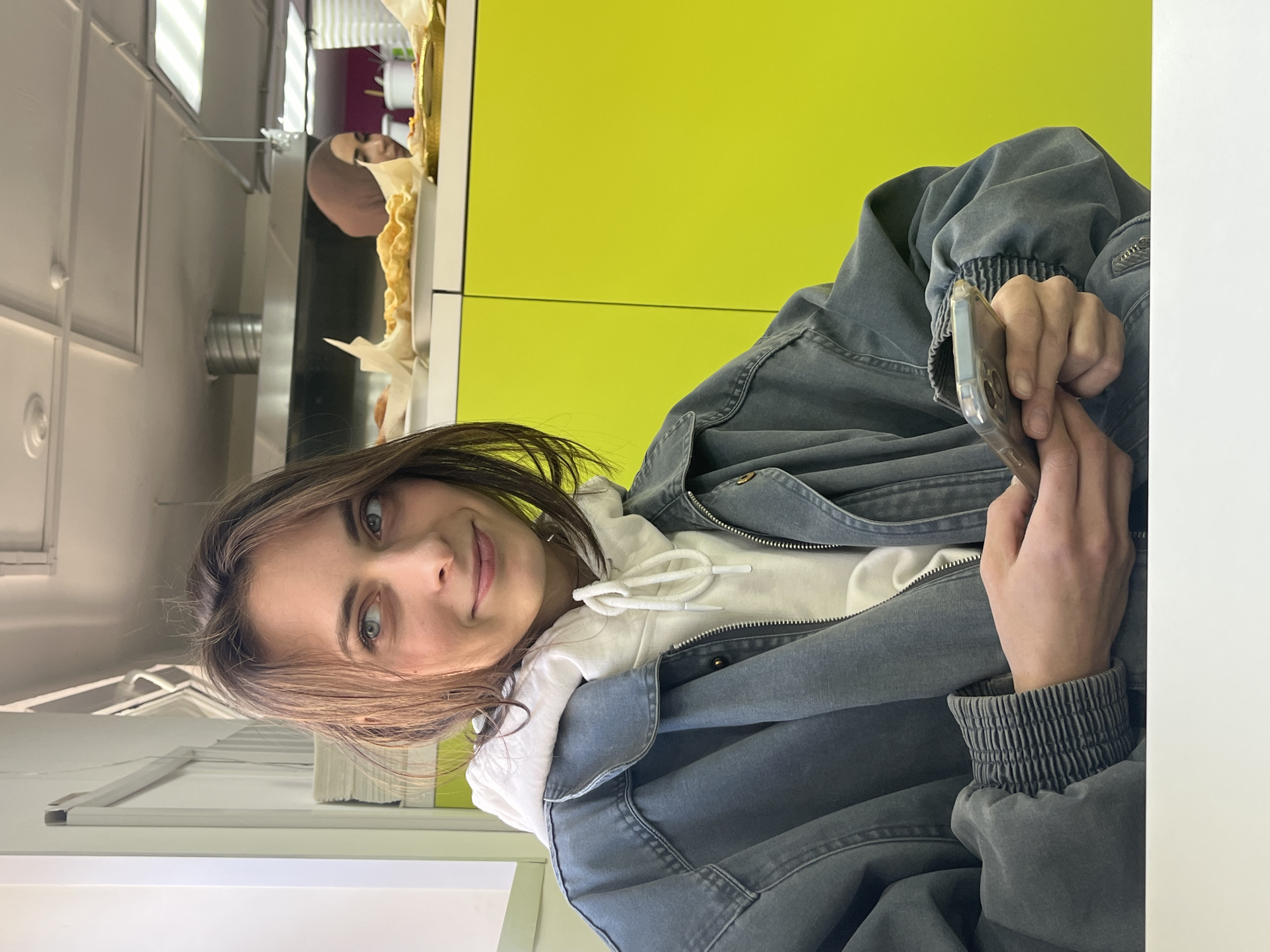 The image, captured from an overhead angle and rotated 90 degrees counter-clockwise, features a young woman as the primary subject, looking directly at the camera with a gentle smile. She has shoulder-length brown hair and striking blue-green eyes. She is wearing a white hooded sweatshirt under a dark grey or faded blue denim-style jacket, which features a zippered front and elasticized, ribbed cuffs. In her right hand, she holds a smartphone encased in a clear protective cover.
The background is dominated by a vibrant, bright yellow wall, divided by a horizontal line, suggesting panels or different sections. To the left of the main subject (when viewed correctly oriented), a second individual is partially visible. This person, likely a woman, is wearing a light brown hijab and appears to be standing behind a counter where items resembling pastries or baked goods, wrapped in white parchment paper, are displayed.
The setting appears to be a modern indoor space, possibly a cafe or cafeteria, characterized by white ceiling panels with integrated lighting fixtures. A white tabletop or counter surface occupies the foreground and parts of the right side of the frame, with stacks of books or papers resting upon it. The lighting is bright and even, indicating an indoor daytime setting. No discernible text is visible within the image. The overall mood conveyed by the main subject is relaxed and friendly.