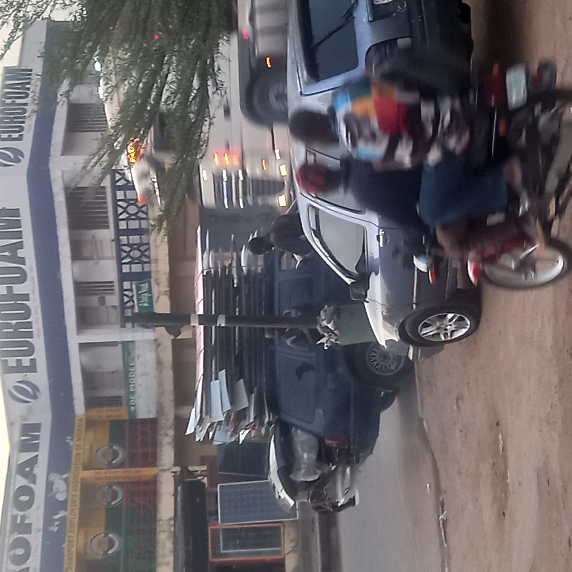 The scene is located in Sardauna, Nigeria. An "EUROFOAM" sign is visible on a building. People on a motorcycle are near a car. There are other vehicles on the road. There are also crates and other objects in front of buildings.