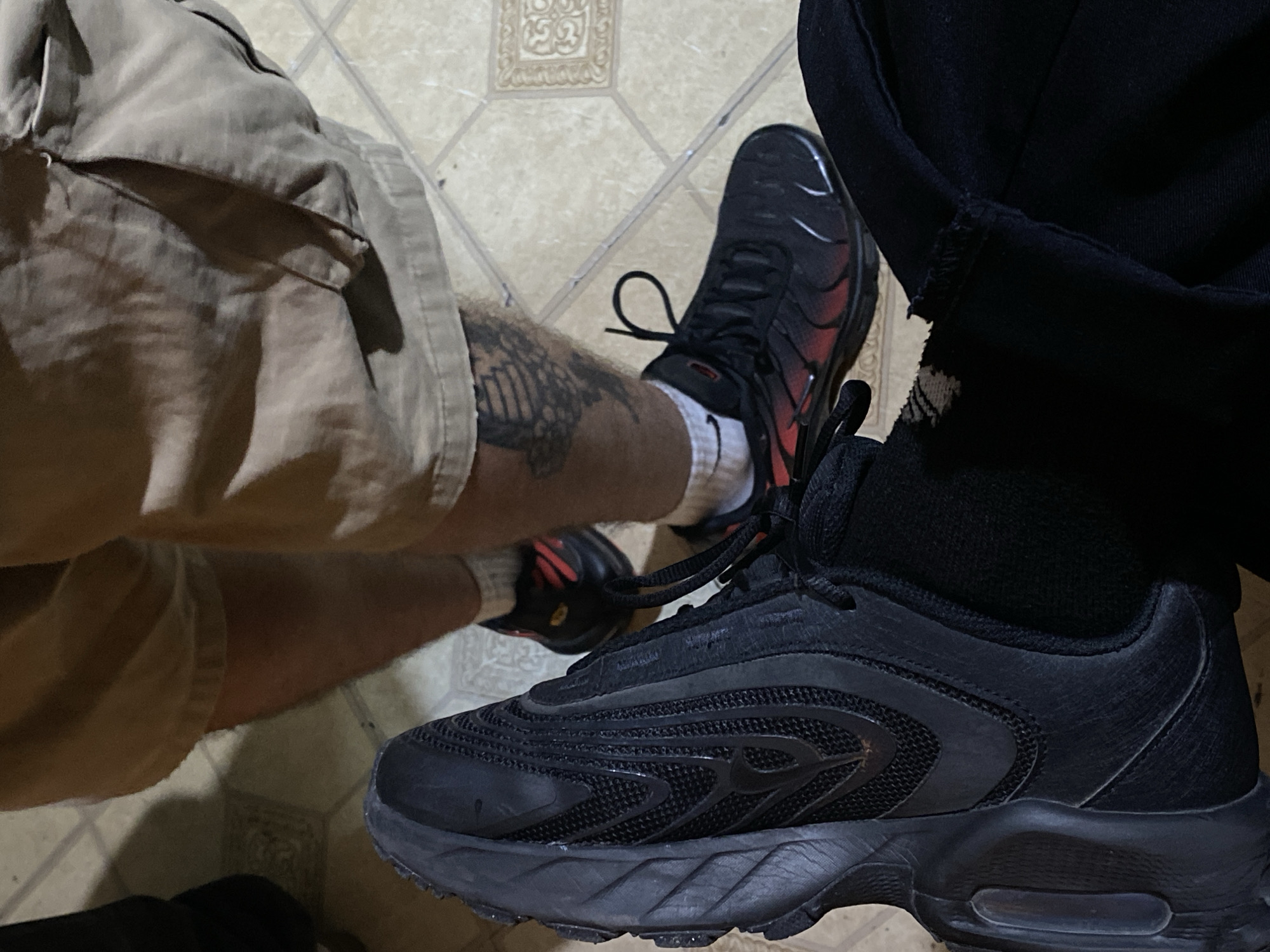The image shows two pairs of legs and feet on a tiled floor. On the left, a person wears khaki shorts, white socks, and black sneakers with red accents. A tattoo is visible on the left leg. On the right, a person wears black pants, black socks, and black sneakers. The location, Monte Grande, Argentina, is not evident from the image.