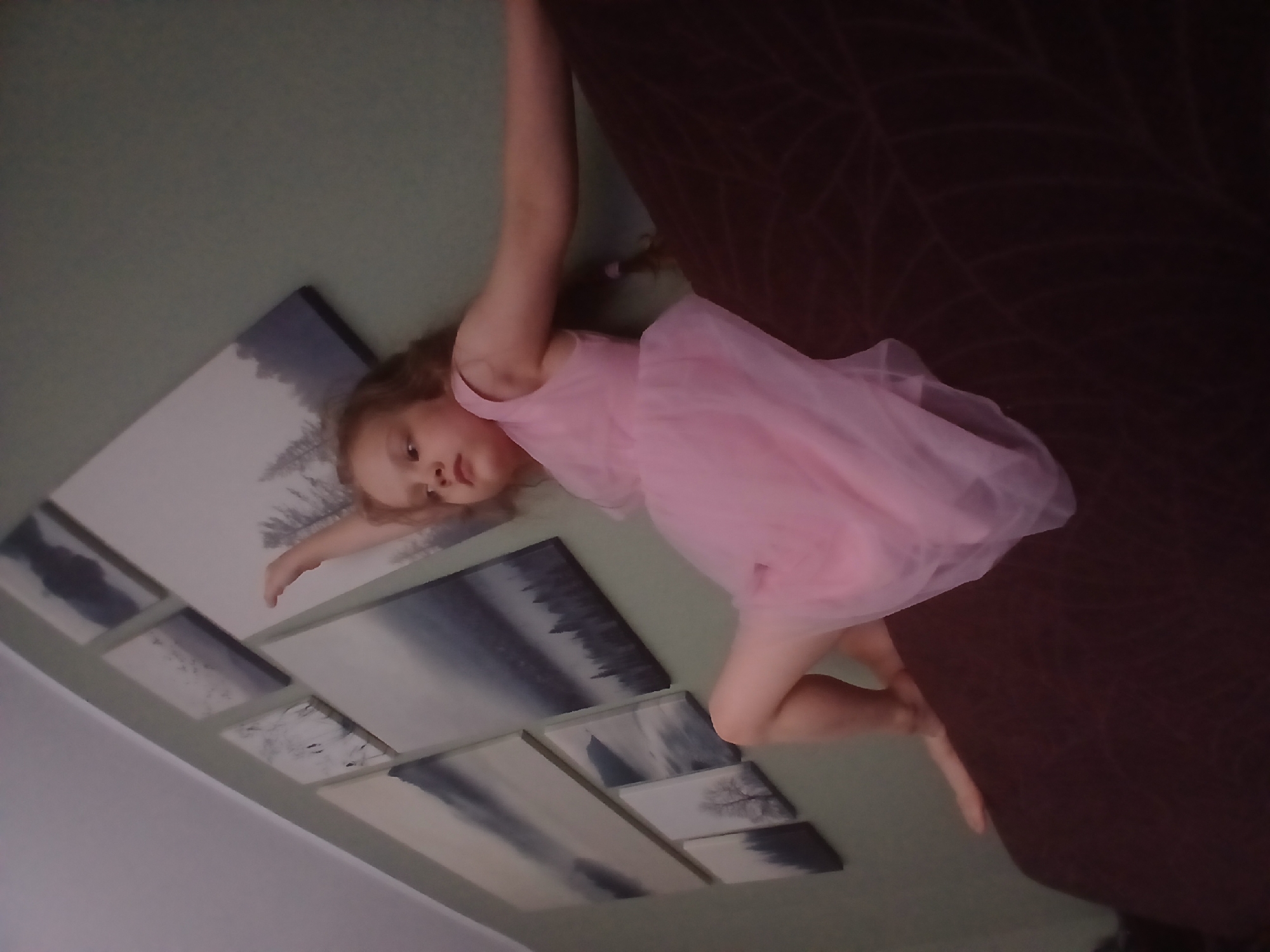 A young girl with blonde hair, wearing a pink dress, is in a reclined position on a dark brown couch. Her right arm is extended outwards, while her left arm is bent at the elbow. Behind her, on a pale green wall, are several framed canvas artworks depicting various landscapes, some featuring trees and water. The overall scene suggests an indoor residential setting. The image does not contain any information that can help determine the location in the city of Yekaterinburg, Russia.