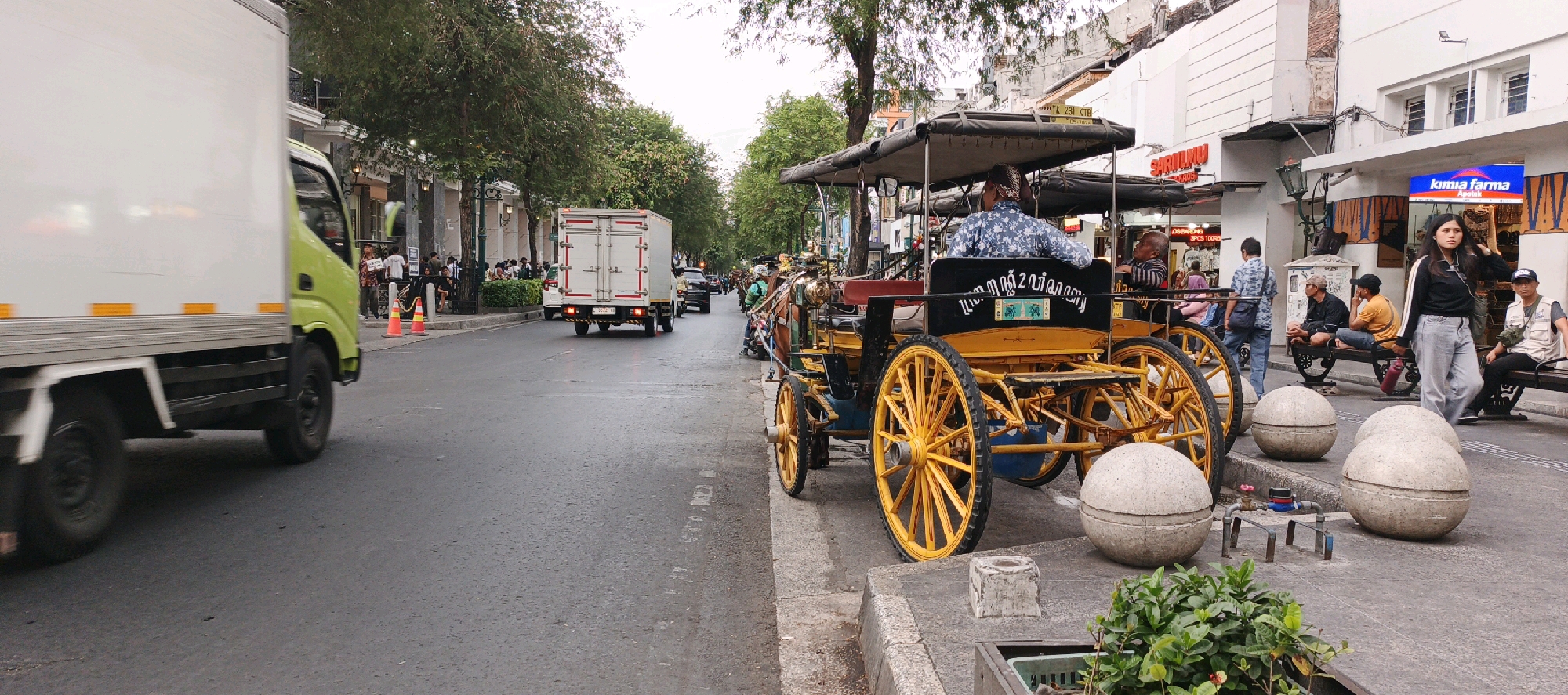This image captures a street scene in Yogyakarta, Indonesia, likely Malioboro Street, a popular tourist destination known for its street vendors, horse-drawn carriages (andongs), and lively atmosphere.
In the foreground, a bright yellow and black horse-drawn carriage, an andong, is prominently featured, its large, ornate wheels suggesting a traditional mode of transport. The carriage is occupied by a man in a patterned blue shirt, presumably the driver, and a passenger, an elderly man wearing a striped shirt, who is looking to his right. Beside the carriage, large, rounded concrete bollards are strategically placed along the sidewalk.
The sidewalk is bustling with activity. A young woman with dark hair is standing, looking to her left, dressed in a black jacket with white stripes and light-colored jeans. To her right, two men are seated on a bench, one wearing a yellow t-shirt and the other a black t-shirt and a cap. Another man in a light vest and cap is seated further to the right on a similar bench. In the background, several other people are visible, some walking and others standing.
The street itself is paved and wet, suggesting recent rain or cleaning. A white delivery truck with a green cab is moving away from the camera on the left side of the road. Further down the street, another white truck is visible, along with other vehicles and more horse-drawn carriages. Buildings line the street on the right, with shops visible, including a sign for "KIMIA FARMA Apotek." Trees with green foliage line the left side of the street, providing shade and adding to the urban landscape. The overall impression is that of a vibrant, active street in a city that blends modern life with traditional elements.