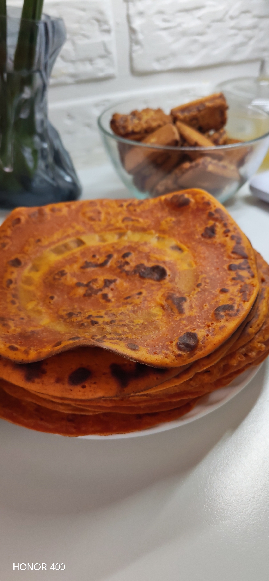 A close-up, slightly angled shot captures a stack of reddish-orange flatbreads or pancakes on a white plate in the foreground. The top flatbread features a textured surface with darker, browned spots and a subtle swirled pattern. In the blurred background, to the right, a clear glass bowl is filled with golden-brown, rectangular baked goods, possibly biscottis or rusks. To the left, a dark, textured vase or decorative object holds faint green elements, likely plant stems. The background wall is white with a textured, brick-like pattern. The setting appears to be an indoor kitchen or dining area, likely prepared for a meal or snack. No people are visible, and therefore no specific emotions are depicted. The time of day is not discernible. The text "HONOR 400" is visible in the bottom left corner of the image.