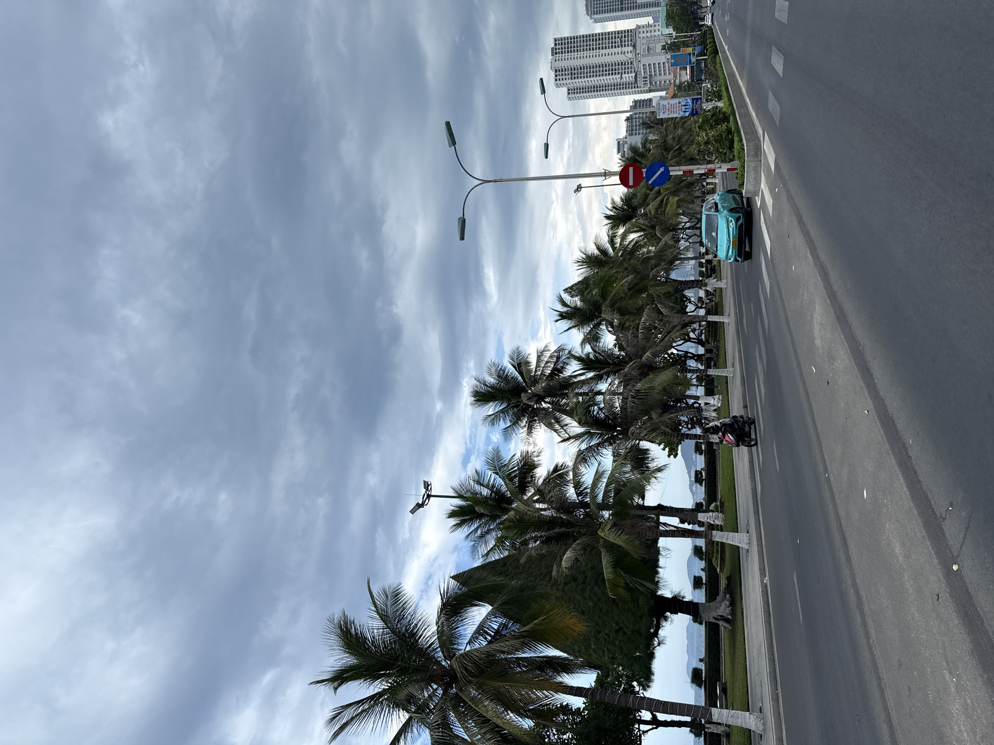The image depicts a street scene in Nha Trang, Vietnam, likely along the beachfront. The sky is overcast with dramatic gray clouds. A row of palm trees lines the left side of the street, with a manicured green space and the ocean visible beyond them. The street itself is a multi-lane asphalt road.
In the foreground, a light blue electric car is stopped at a crosswalk, facing the viewer's right. A traffic sign with a red circle and a blue arrow is visible next to the car. Further down the road, a person on a red scooter is riding away from the viewer, also crossing the street.
In the background, several tall, modern buildings dominate the skyline, suggesting an urban coastal environment. The overall impression is a somewhat subdued day in a bustling coastal city.