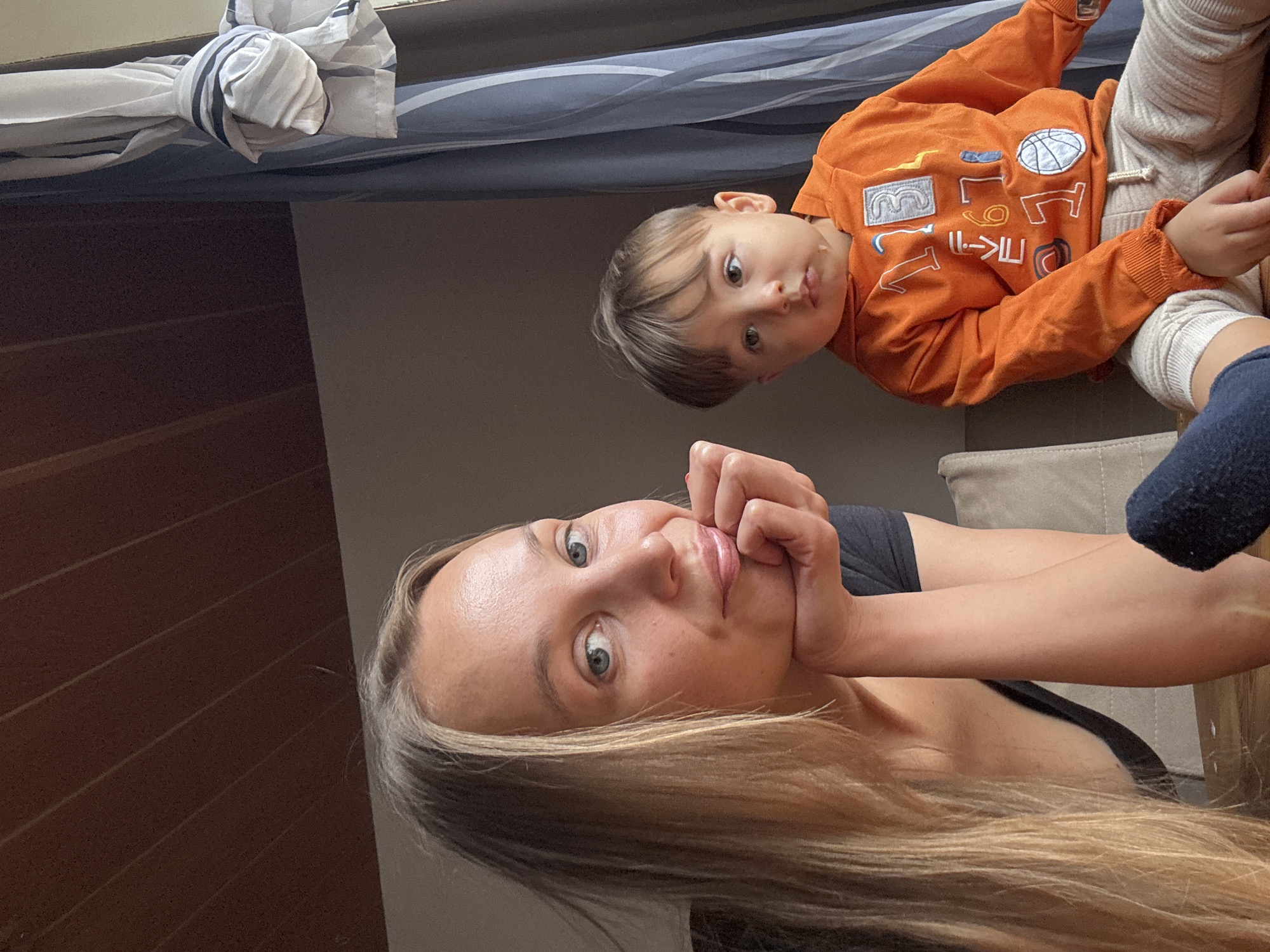 A woman with long blonde hair is in the foreground, resting her chin on her hand and looking towards the camera. To her right, a young child in an orange hoodie and light-colored pants is seated, looking away from the camera. The background features a wooden ceiling, a curtain, and what appears to be a window. The scene is indoors, likely in a residential setting. There is no indication of the specific location within Überherrn, Germany, or any public space.