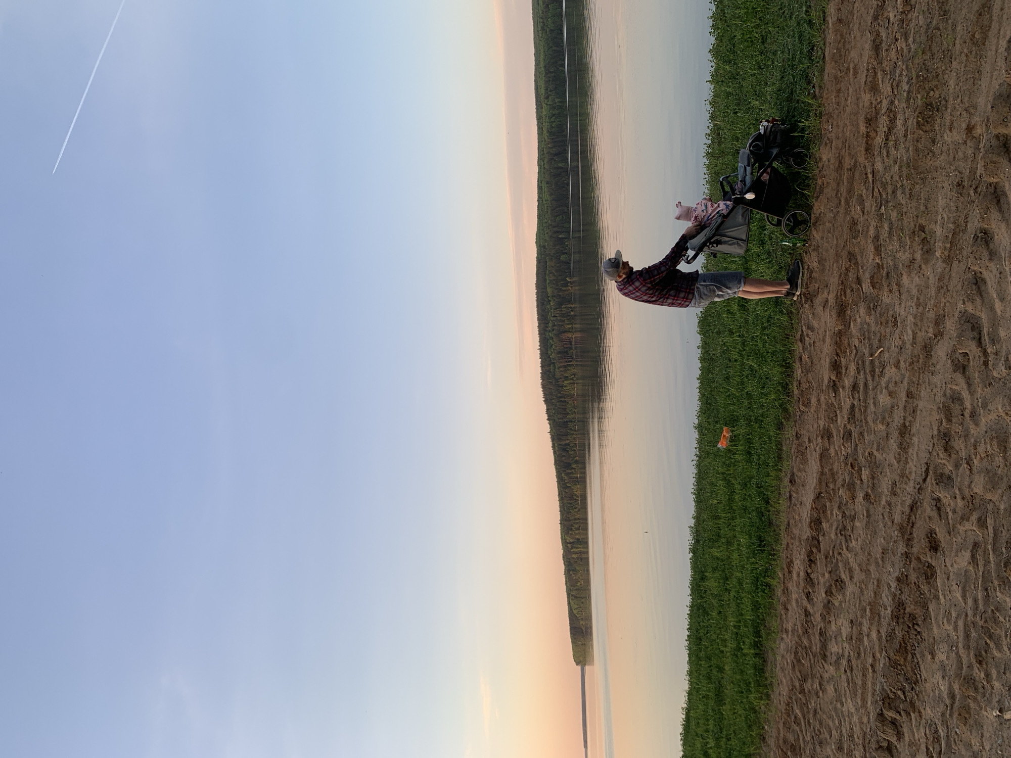 A man is standing on a sandy/dirt embankment next to a body of water. The man is wearing a red and black plaid shirt, shorts, a baseball cap, and dark sneakers. He is pushing a stroller with a baby inside. The water appears calm, with a reflection of a treeline on the opposite bank. The sky is a light blue gradient, with a contrail visible in the upper left. The location is likely Chaikovsky, Russia.