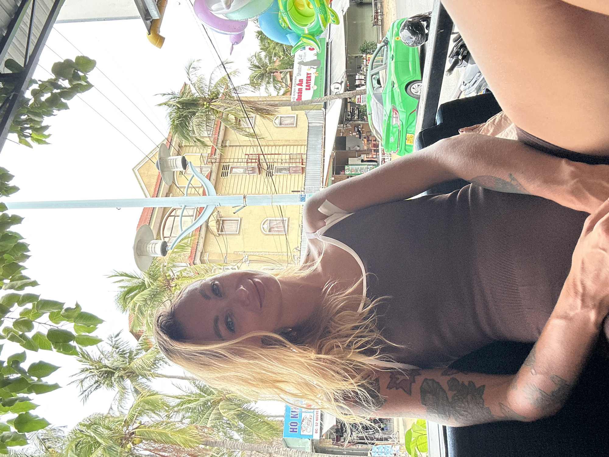 A Caucasian woman with blonde hair, wearing a brown ribbed tank top, is seated in a dark vehicle, looking at the camera with a slight smile. She has tattoos on her left arm and what appears to be a bandage or medical tape on her right upper arm. The background shows a street scene in Phu Thuy Ward, Vietnam, with palm trees, a yellow building with red balconies, and a bright green car. Other elements in the background include shops, signs with Vietnamese text, power lines, and colorful inflatable decorations. The overall lighting suggests it is daytime.