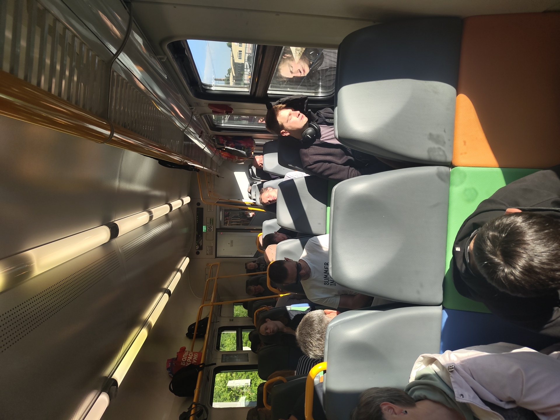 The image depicts the interior of a train carriage, viewed from the perspective of a seated passenger. Sunlight streams in through the windows, illuminating the aisle and casting long shadows. Several passengers are visible, some sleeping, others looking out the window or towards the front of the train. The seats are upholstered in grey with green and orange fabric accents on the lower sections. Luggage is stored on overhead racks. The train appears to be moving through a rural or suburban area, as indicated by the greenery visible through the windows. The number "06" is visible on a sign above a doorway. The scene is consistent with a commuter or regional train in Russia.