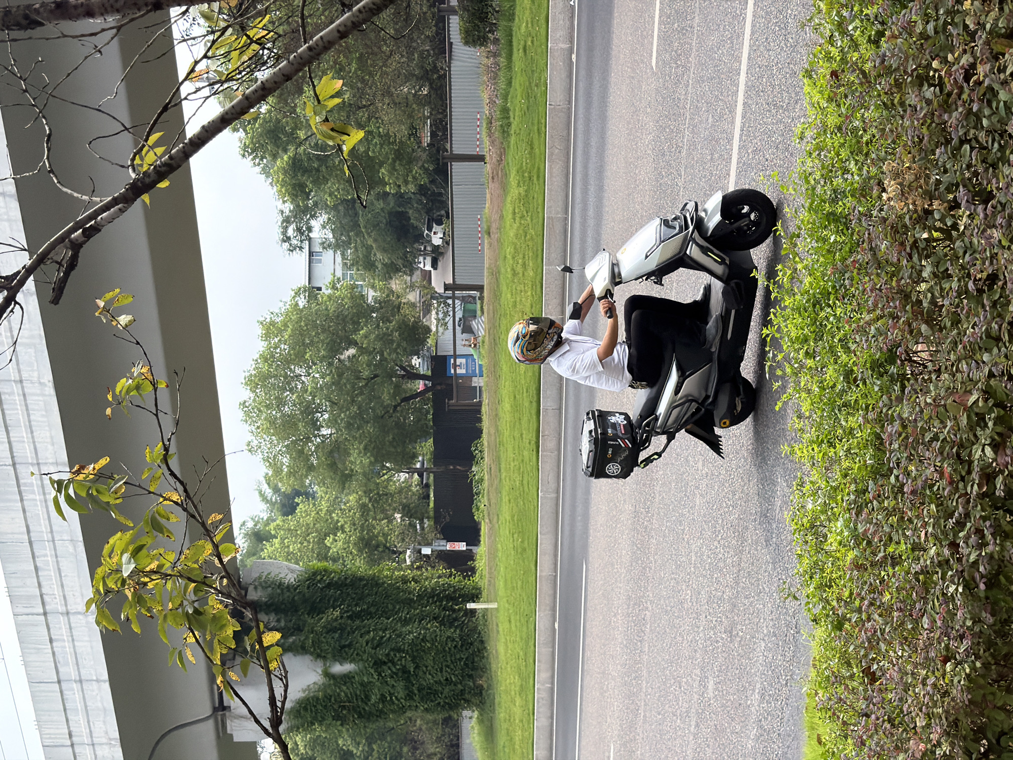 A person wearing a white short-sleeved shirt, black pants, and a helmet rides a silver electric scooter on an asphalt road in Jiangjin, China. The scooter has a black storage box mounted on the rear. The road is bordered by a grassy median and a sidewalk, beyond which are trees and a building. An elevated highway structure is visible in the background, with vines growing on a concrete support. The image is taken from a slightly elevated perspective, looking down at the scooter.