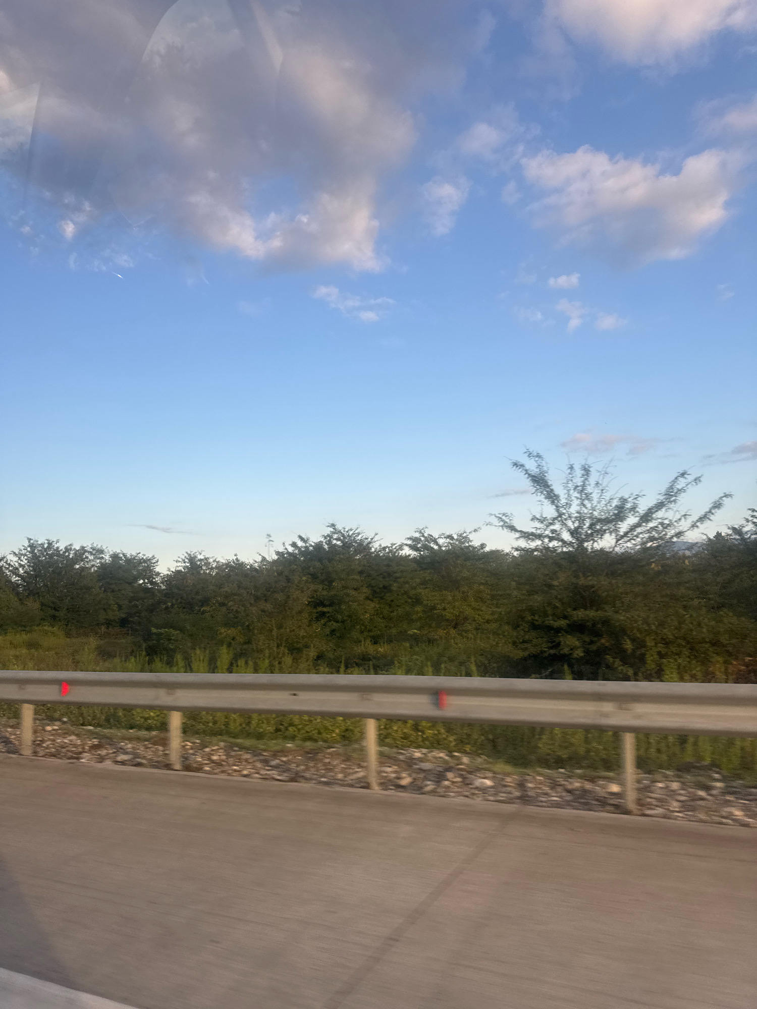 A photograph taken from a moving vehicle captures a roadside scene in Ukaneti, Georgia. A grey concrete road occupies the foreground, with a reflective metal guardrail running parallel to it. Beyond the guardrail, a dense line of green trees forms a natural barrier, partially obscuring a distant, hazy landscape. The sky above is a clear blue, with a scattering of white and grey clouds. The overall impression is one of a rural or semi-rural environment, possibly on the outskirts of Ukaneti.