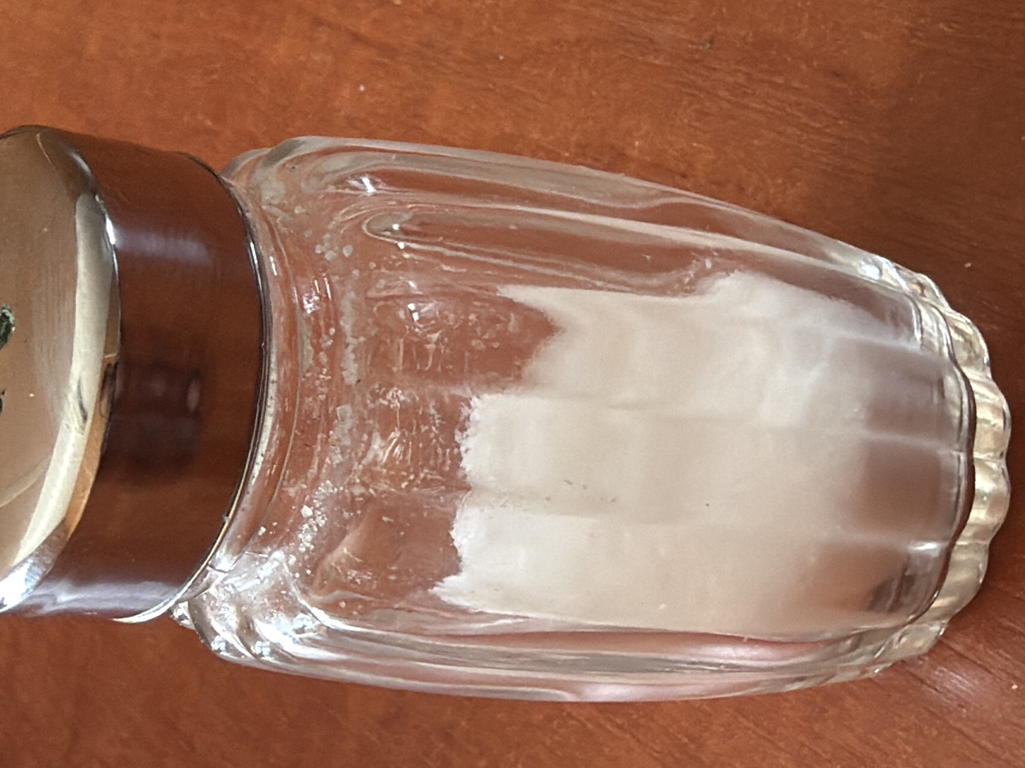 A close-up, high-angle shot shows a glass salt shaker filled with white granules, resting on a textured brown surface, likely a wooden table. The salt shaker has a fluted glass body and a metallic, possibly chrome, lid. The salt inside is clumped in some areas, suggesting it may have been exposed to moisture. The lighting is somewhat dim, casting subtle shadows. There are no people or any other objects visible in the frame. The scene is devoid of any context that would suggest a specific location within Janowice, Poland.