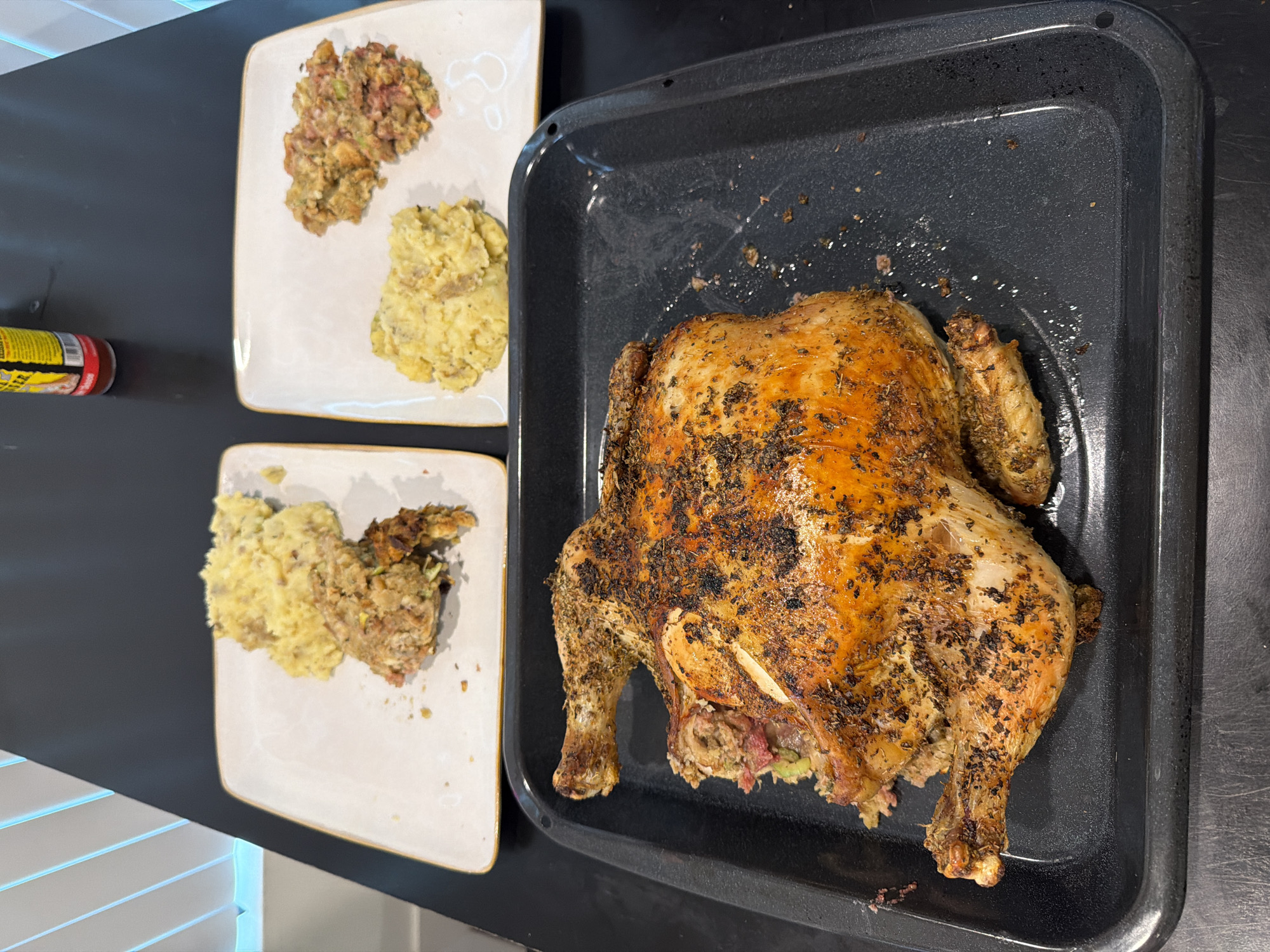 The image presents a roasted chicken on a black baking tray, accompanied by two plates containing portions of mashed potatoes and stuffing. The setting appears to be a domestic kitchen, inferred from the countertop surface and the presence of what looks like a jar of condiments. The scene is staged for a meal, showcasing a complete dish ready for serving. The absence of people and any immediate context prevents specifying a specific location in Los Angeles.