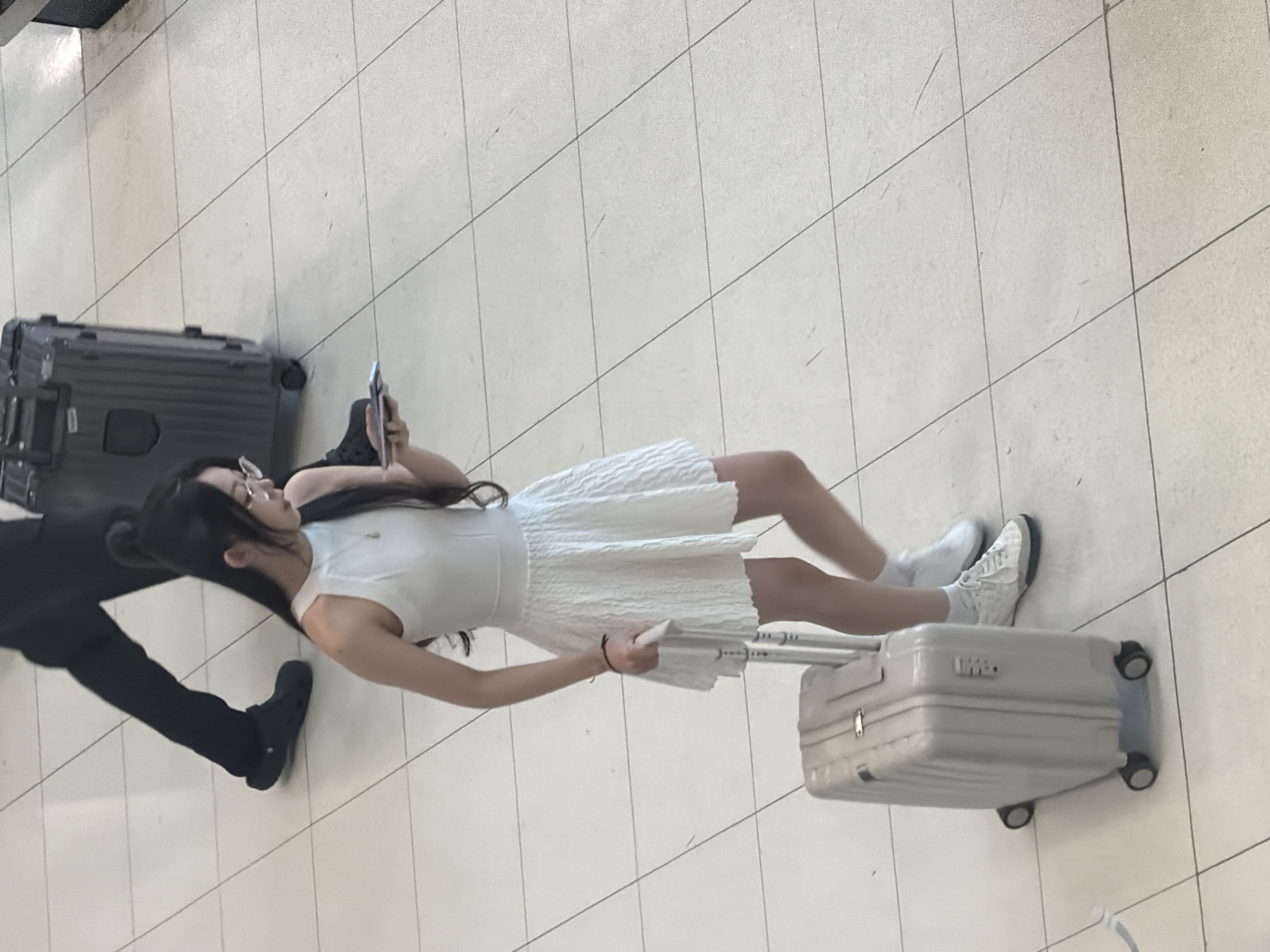 A young woman with long, dark hair tied in a bun and wearing glasses, a sleeveless white dress, and white sneakers is standing in what appears to be an airport terminal. She is holding a mobile phone and a passport in her hands. Beside her, she is pulling a light-colored suitcase with a retractable handle. In the background, another person is walking away, wearing dark pants and dark shoes. The floor is tiled with large, light-colored square tiles. The scene is captured from a slightly elevated angle. The location is identified as Nong Prue Subdistrict, Thailand.
