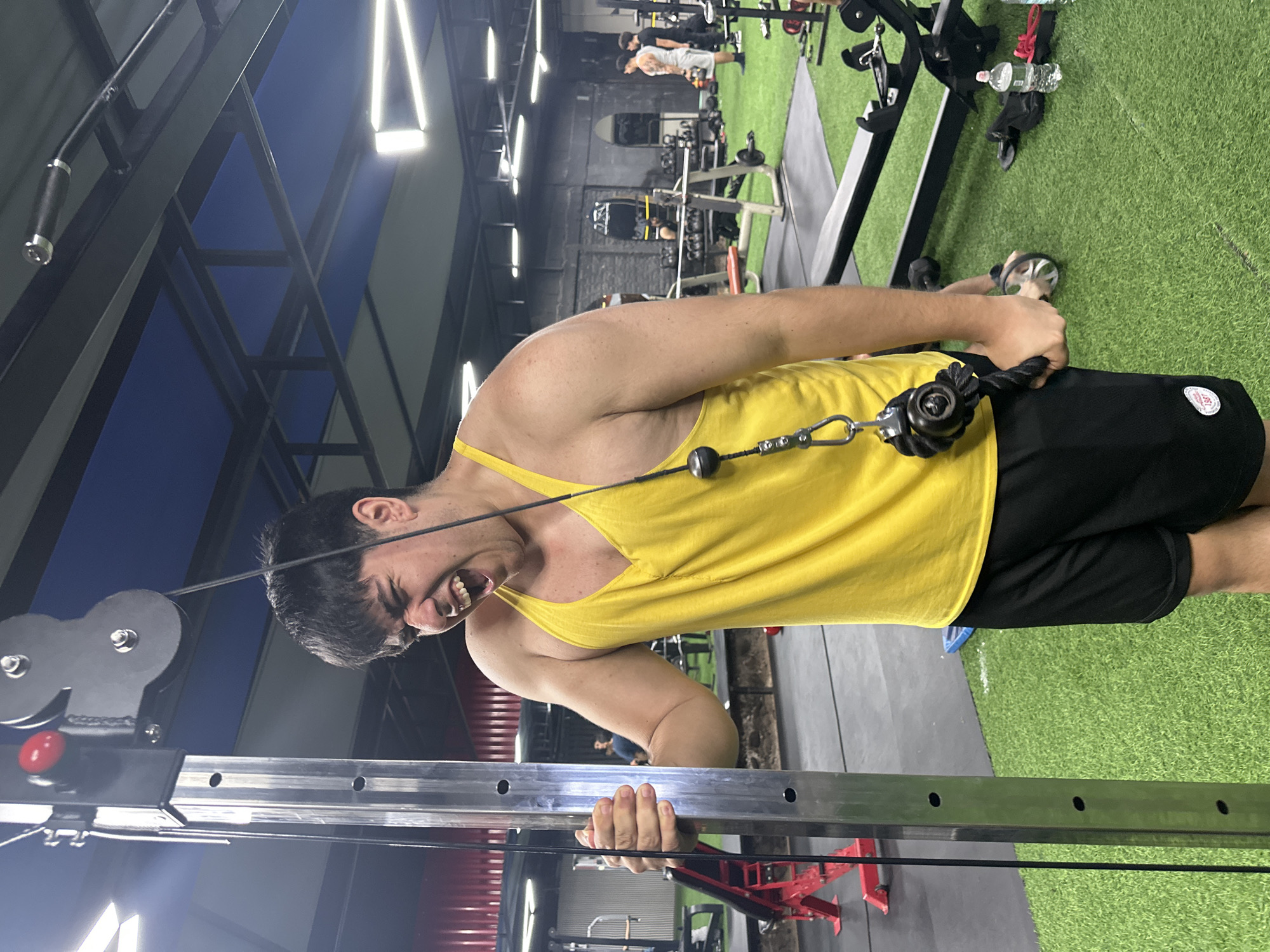 A person in a yellow tank top and black shorts is using a cable machine in a gym. The person is grimacing, suggesting exertion. The gym has artificial turf flooring and various exercise equipment. The location appears to be indoors, possibly in Arica, Chile.