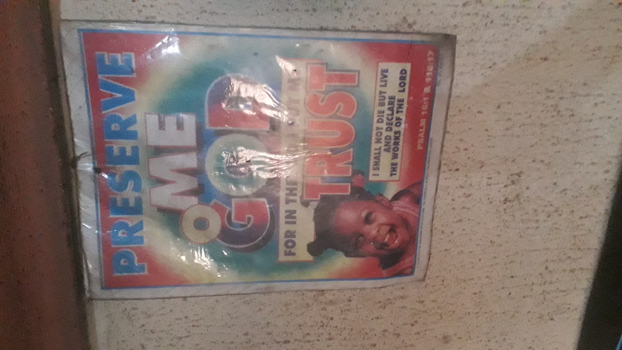 A weathered, plastic-covered poster is affixed to a rough, textured wall in Nkpor, Nigeria. The poster features a vibrant, abstract background with colors transitioning from red at the top to blue at the bottom, with green and yellow in between. Dominating the poster are large, bold letters spelling out "PRESERVE ME GOD TRUST." Below the large text, a smiling baby with dark skin and hair styled in two small buns is pictured on the left side. To the right of the baby, a biblical quote reads, "I SHALL NOT DIE BUT LIVE AND DECLARE THE WORKS OF THE LORD." Below this quote, smaller text indicates "PSALM 16:1 & 116:17." The poster appears to be aged, with some creasing and a slight sheen from the plastic covering, suggesting it has been exposed to the elements. The wall itself is a light, mottled beige with visible speckles and texture, indicative of a typical outdoor or semi-outdoor environment in a Nigerian urban setting.