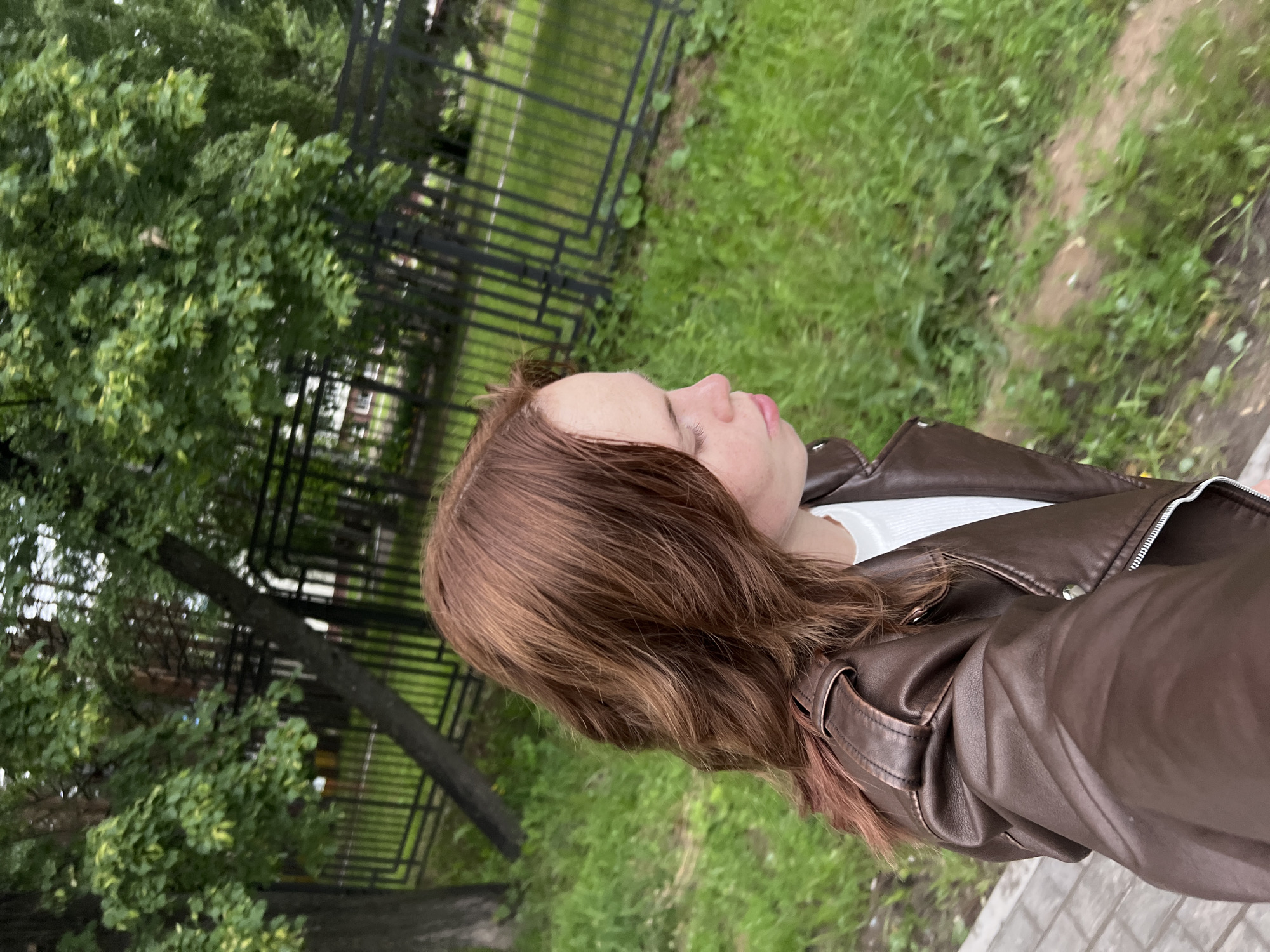 The image depicts an outdoor scene in the city of Chaikovsky, Russia, featuring a young female from the chest up, viewed in profile. She has reddish-brown, wavy hair that extends past her shoulders. She is wearing a dark brown leather jacket with shoulder epaulets and a silver zipper, over a white ribbed top. Her face is turned to the right, with her eyes appearing closed or downcast, and her lips slightly pursed. She is positioned on the left side of the frame, standing on a paved walkway comprised of light-colored rectangular pavers. To the right of the walkway is a grassy area with some patches of bare earth. In the mid-ground, a black metal fence with vertical bars and horizontal rails runs across the frame, separating the foreground from a background dominated by dense green foliage from numerous trees. The lighting suggests an overcast or soft light condition.