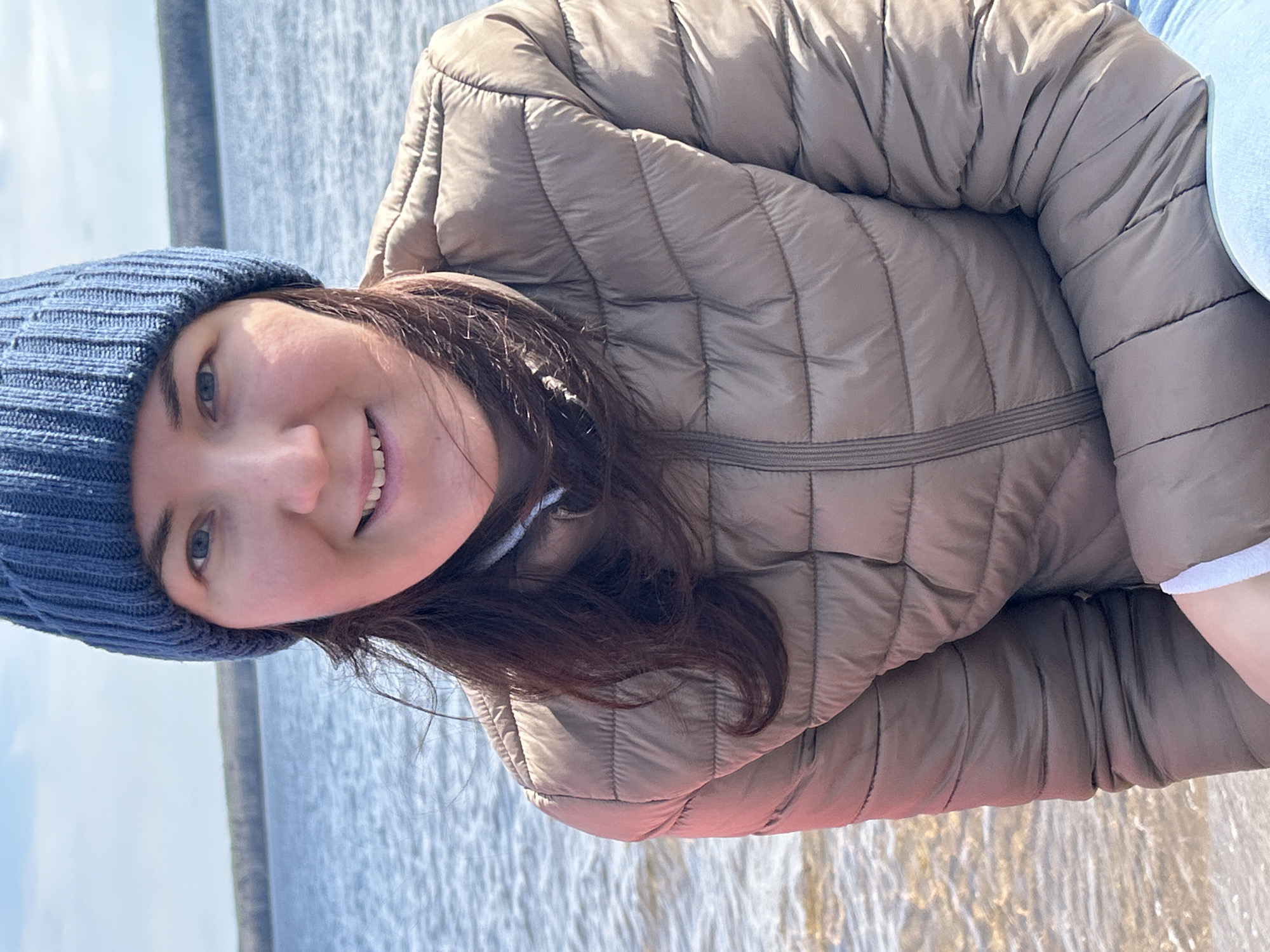 A young woman is captured in a vibrant outdoor portrait, smiling brightly directly at the camera. Her head is tilted slightly, and her dark brown hair falls loosely around her shoulders, partially covered by a ribbed blue knitted beanie. She has blue eyes and a wide, cheerful smile, revealing her teeth. She is wearing a beige or light brown puffer jacket with horizontal quilting, suggesting the weather is cool or cold.
The background features a large body of water, likely a lake or river, under a bright, partly cloudy sky. The water's surface shows subtle ripples, indicating a calm environment. A faint horizon line with distant land can be seen across the water. The scene appears to be set during the daytime, with ample natural light illuminating the subject and the landscape. The woman's attire and the natural setting are consistent with an outdoor activity or casual outing in a location like Chaikovsky, Russia, potentially near the Votkinsk Reservoir or the Kama River. There is no visible text in the image.