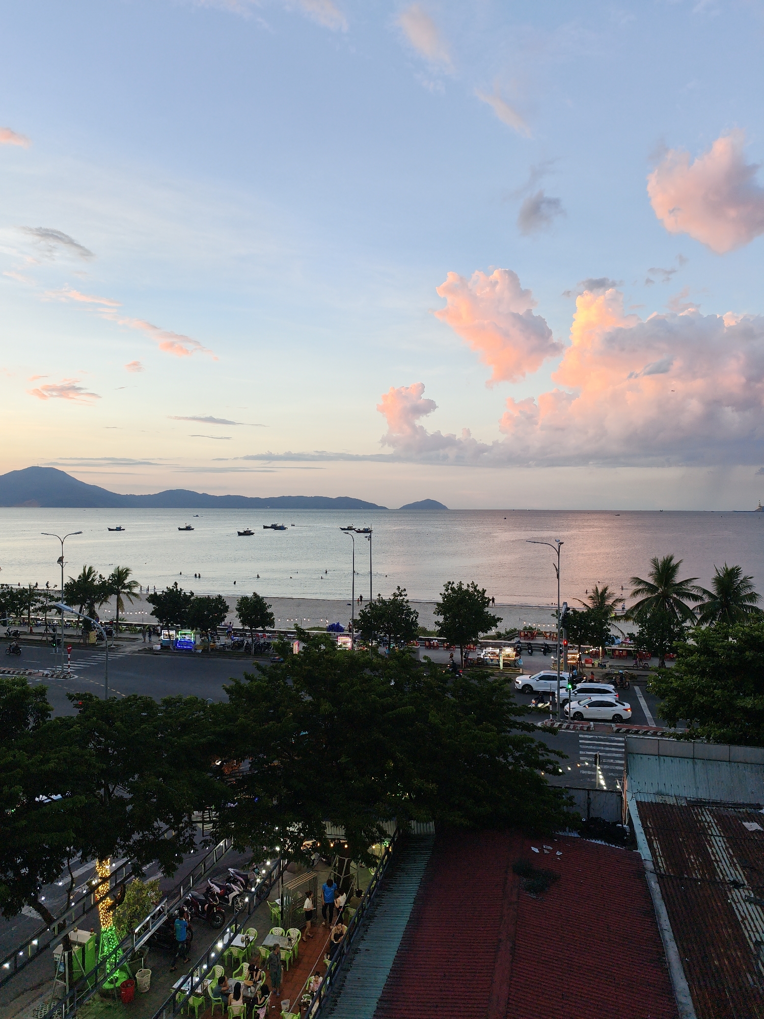 The image depicts a panoramic view of a beachside city, likely Da Nang, Vietnam, during the late afternoon or early evening. The sky is a blend of soft blues and pastel pinks and oranges from the setting sun, with scattered clouds reflecting the warm light. The ocean stretches to the horizon, dotted with several small boats. A wide sandy beach is visible along the coastline, with a few people scattered along its expanse.
In the foreground, a street runs parallel to the beach, with cars and motorcycles navigating the road. Trees and palm trees line the street, providing shade. Further in the foreground, an outdoor dining area is set up with tables and chairs, illuminated by string lights. People are seated at the tables, some appear to be dining, while others are socializing. The scene suggests a vibrant, active coastal city scene, with leisure and transport coexisting.