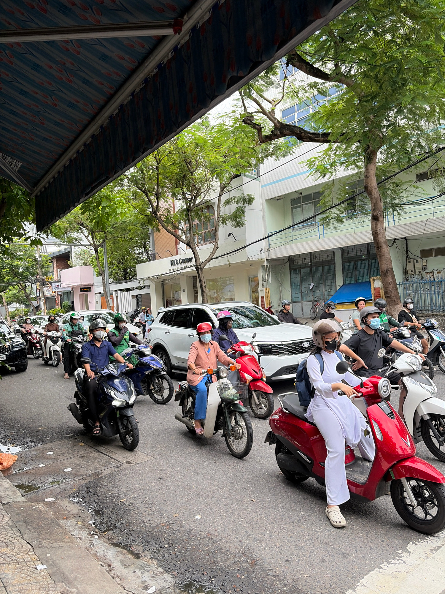 This is a street-level, eye-level, medium shot of a busy street scene in Da Nang, Vietnam. The perspective is from behind a street awning, looking down and across the road. The street is lined with buildings on the right, some with balconies and windows, and trees overhang the street from the left. A group of people on motorbikes are moving through traffic.
In the foreground, a woman in a white traditional Vietnamese outfit and a white helmet is riding a red scooter. She is wearing white clogs. To her left, a person on a dark blue scooter wears a dark blue shirt and helmet, and their face is partially obscured by a mask. Behind them, a person on a green and black scooter wears a green shirt and helmet. Further back, a white SUV is visible, followed by several other motorbikes. On the left side of the image, more motorbikes and people are seen in traffic, including a black car. The road surface is asphalt and appears somewhat worn, with some puddles and debris visible. The overall atmosphere is one of daily urban life and transportation in Vietnam.