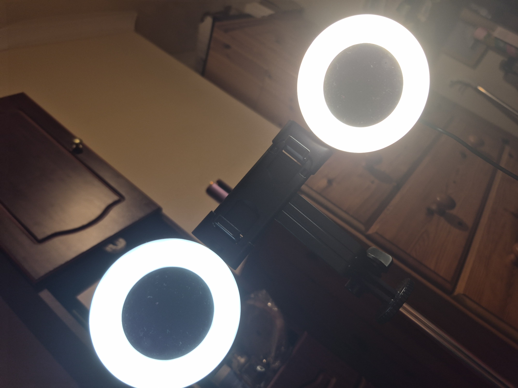 The image shows two ring lights, one at the top left and another at the bottom right, attached to a black stand with a clamp. The ring lights are illuminated with a bright white light. The background consists of wooden furniture, likely drawers and cabinets, with brass handles. The lighting is artificial and bright, coming from the ring lights. The overall environment appears to be an indoor setting, possibly a room or studio. There is no visible indication of a specific location within the city of Sandwell, United Kingdom. No people or interactions are depicted.