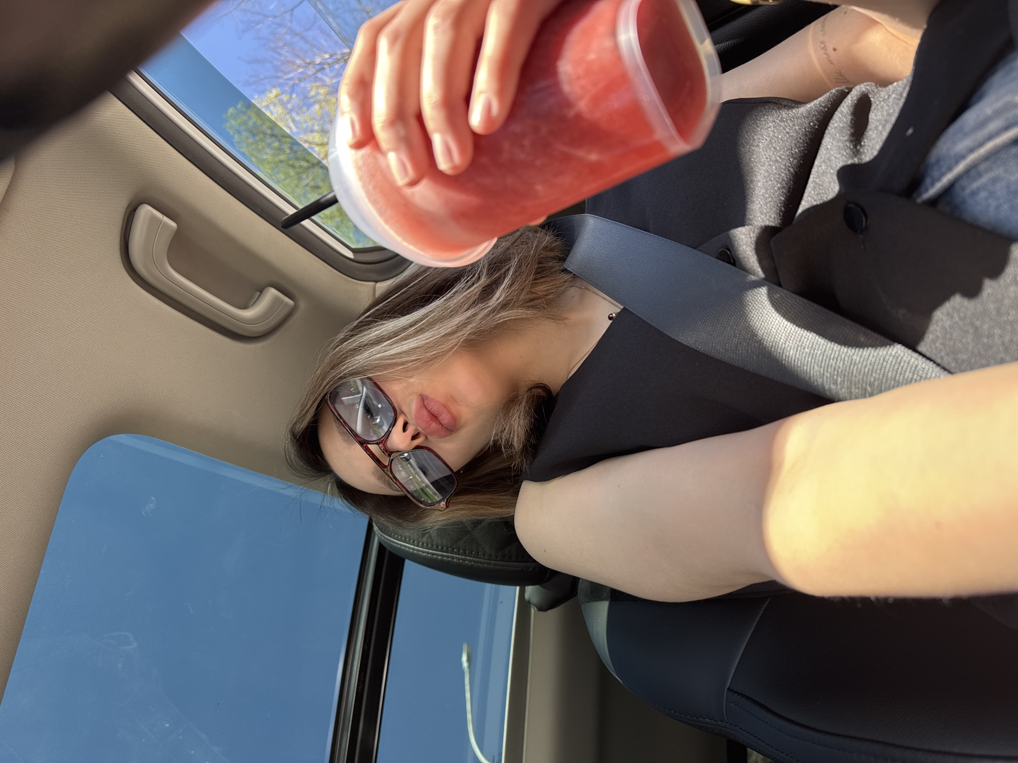 A person wearing glasses and a seatbelt is holding a red beverage in a car, with the sunroof open. The environment shows a blue sky and trees through the sunroof. The location is unknown.