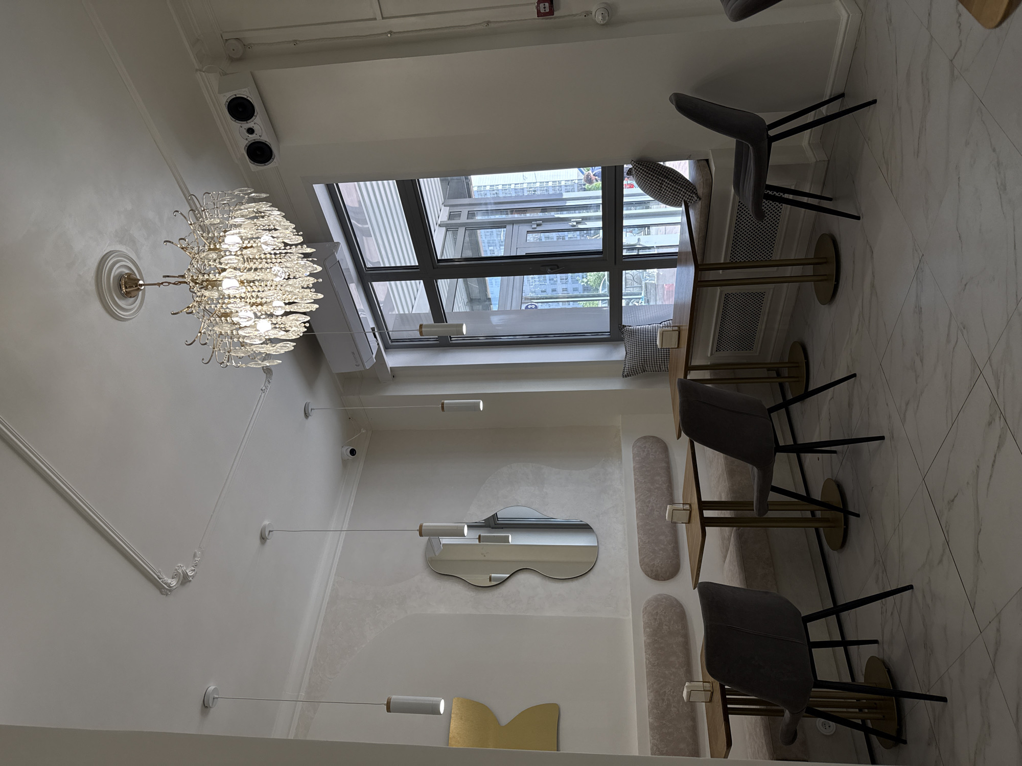 The image displays the bright and modern interior of what appears to be a cafe or restaurant, possibly located in Krasnoyarsk, Russia. The space features white walls, a white marble-patterned tiled floor, and a mix of contemporary and classic design elements.
On the left, a large, elaborate crystal chandelier with multiple lights hangs from the ceiling, affixed to a decorative medallion. Above it, a white rectangular speaker unit is mounted near the ceiling, along with a smoke detector and a fire alarm. Further along the wall, decorative white moldings trace the ceiling line.
The central part of the room is dominated by a large, multi-paned window, offering a view of city buildings and possibly some greenery outside. Below the window, an air conditioning unit is visible. On the wall to the left of the window, an irregularly shaped mirror reflects parts of the interior. Two cylindrical pendant lights hang from the ceiling in this area, with one appearing to be reflected in the mirror. A golden, 'M'-shaped decorative element is affixed to the lower wall.
To the right, dining arrangements consist of square tables with golden bases and wooden tops, paired with dark gray upholstered chairs that have black legs. These tables are set against a built-in sofa-like bench, which is accented by vertically elongated, oval upholstered panels on the wall. Small checkered throw pillows are placed on the bench seating. Additional ceiling-mounted security cameras are visible.
The overall ambiance is clean, well-lit by both natural light from the window and artificial lighting from the chandelier and pendant lights, suggesting a welcoming environment. There are no people visible in the scene, and no discernible text can be read anywhere in the image.