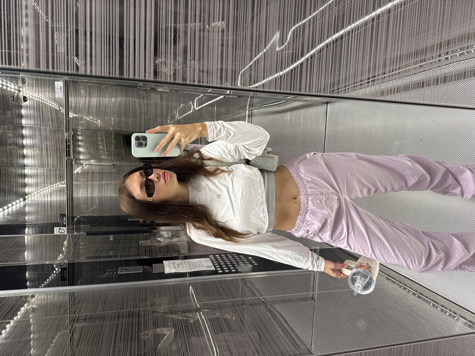 A woman takes a mirror selfie in an elevator. She is wearing sunglasses, a white long-sleeved shirt, and lavender pants. She is holding a phone in one hand and a cup with a straw in the other. The elevator interior has mirrored walls and a metal mesh design. The elevator is in the city of Tel-Aviv, Israel.