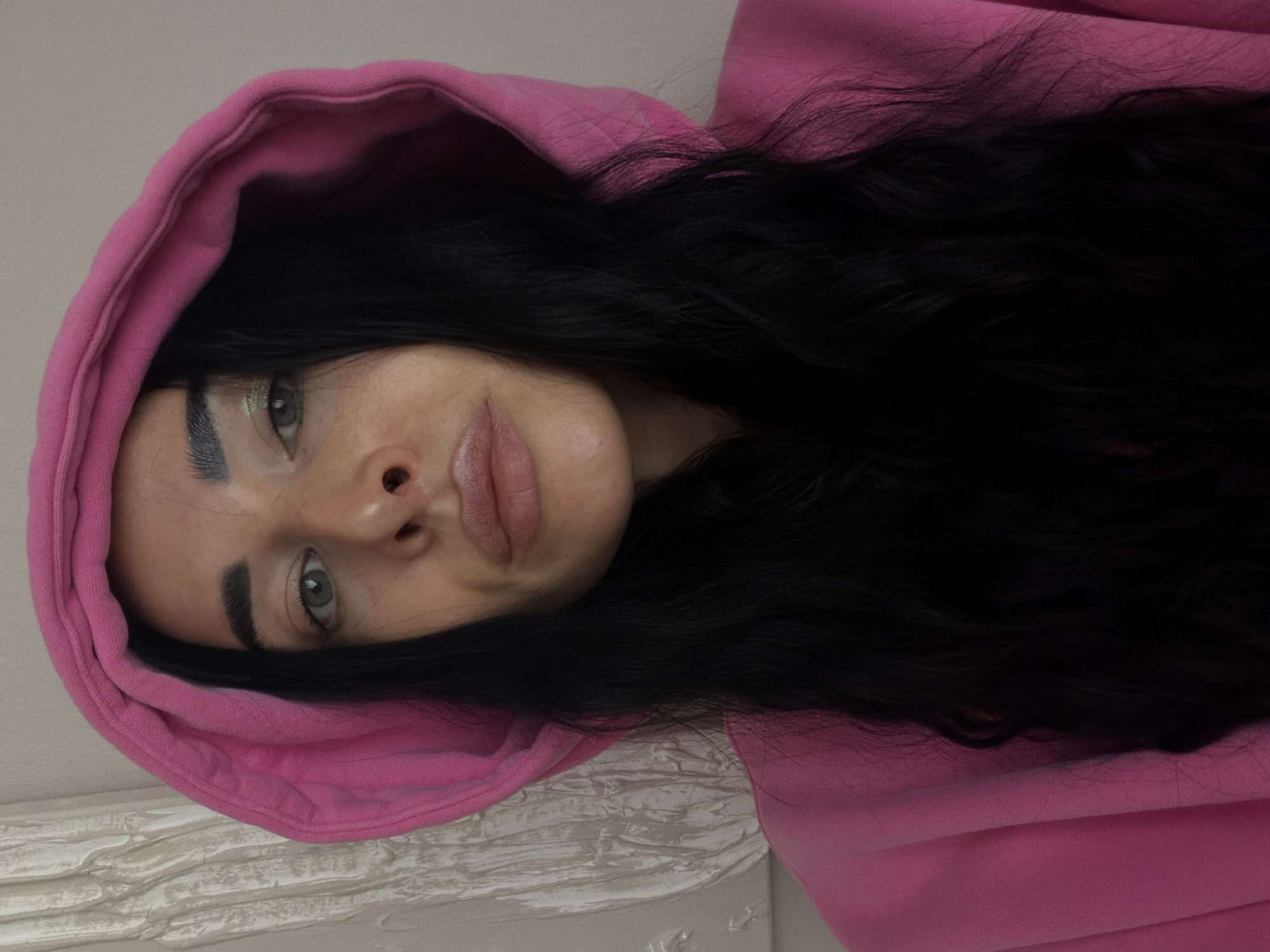 A woman is photographed from an unusual angle, positioned on her side with her head tilted upward. She wears a bright pink hooded sweatshirt, the hood framing her face and partly obscuring her black, wavy hair. Her makeup is subtle, with a slight shimmer visible on her eyelids and gloss on her lips. Her eyes are light, possibly green or grey. The background appears to be a neutral-colored wall with a textured surface, possibly stucco or plaster. The photo does not show any specific details indicating its location in Saint Petersburg, Russia, or any other geographical location.