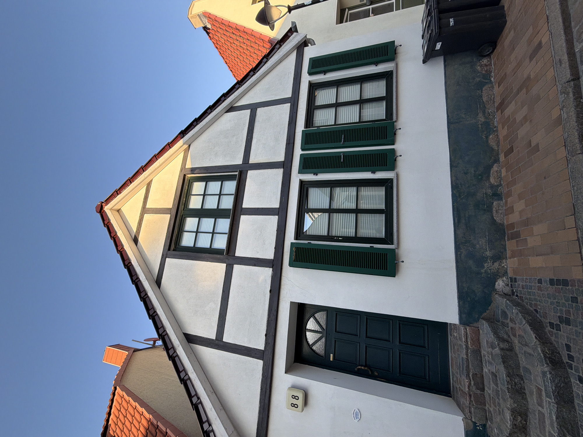 The image shows a close-up, low-angle exterior shot of a historic half-timbered building located in Rostock, Germany. The building features white stucco walls with dark timber framing, a red-tiled roof, and green shutters on its windows. A dark green door with a semicircular glass pane is visible on the left side of the facade, above a set of stone steps. Above the door, the house number "88" is displayed on a white plaque. The street in front of the house is paved with bricks. The sky is clear and blue, indicating fair weather. There are no people or animals visible in the frame. The scene is devoid of any activity, presenting a static architectural view.